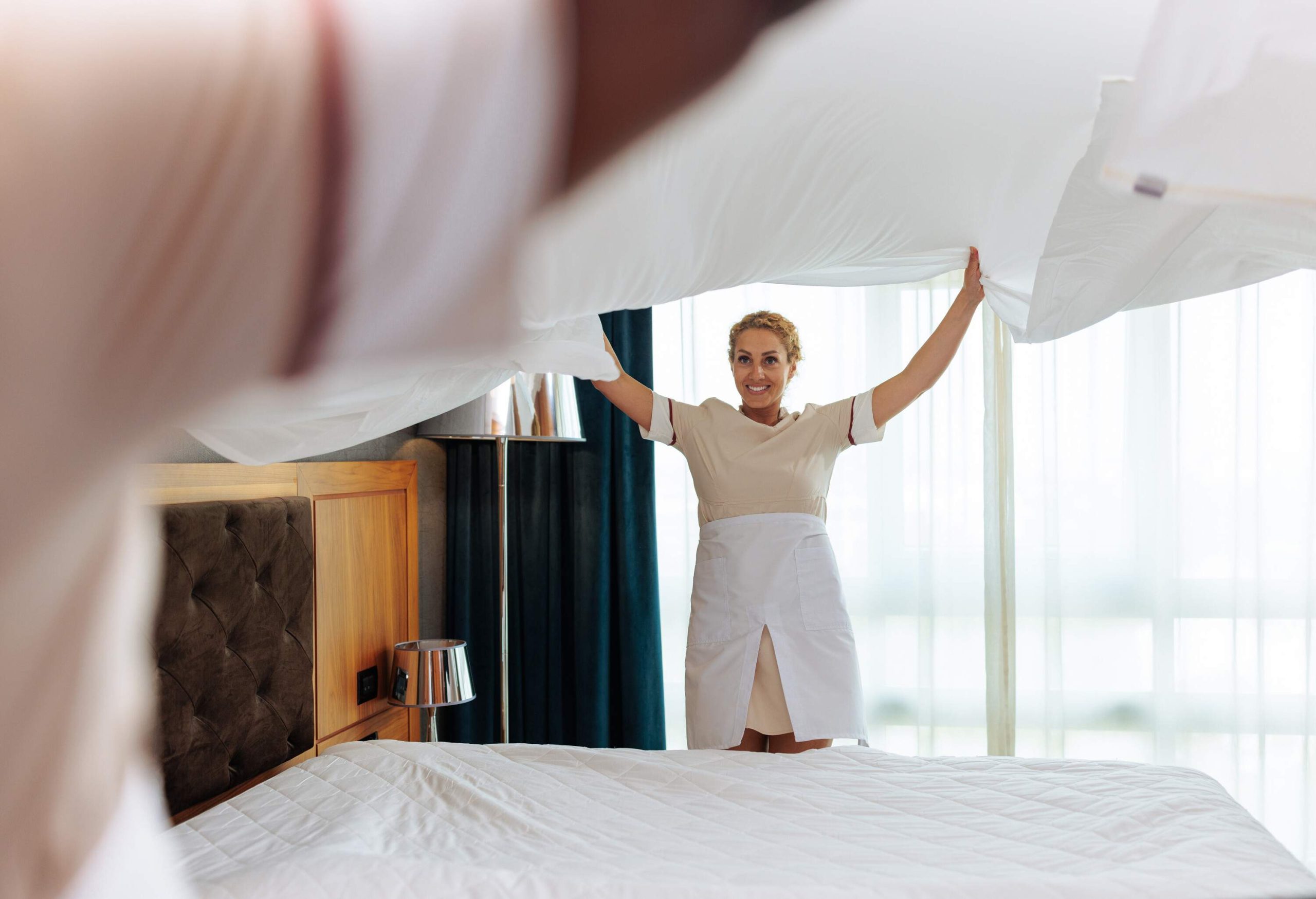 Two young multiracial hotel maids spreading the clean bed sheet high in the air together