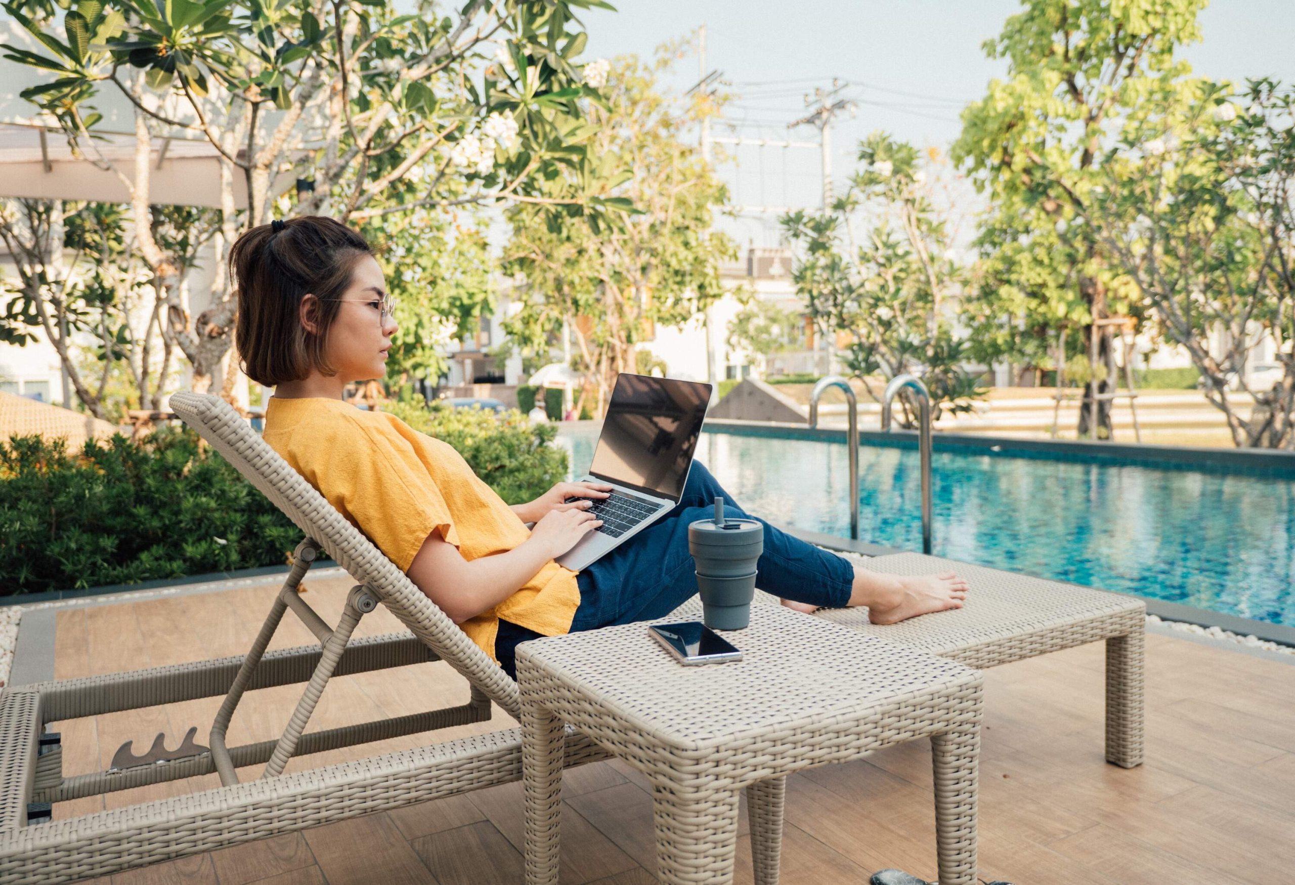 A young woman sitting on a lounge chair by the pool with her laptop on her lap.