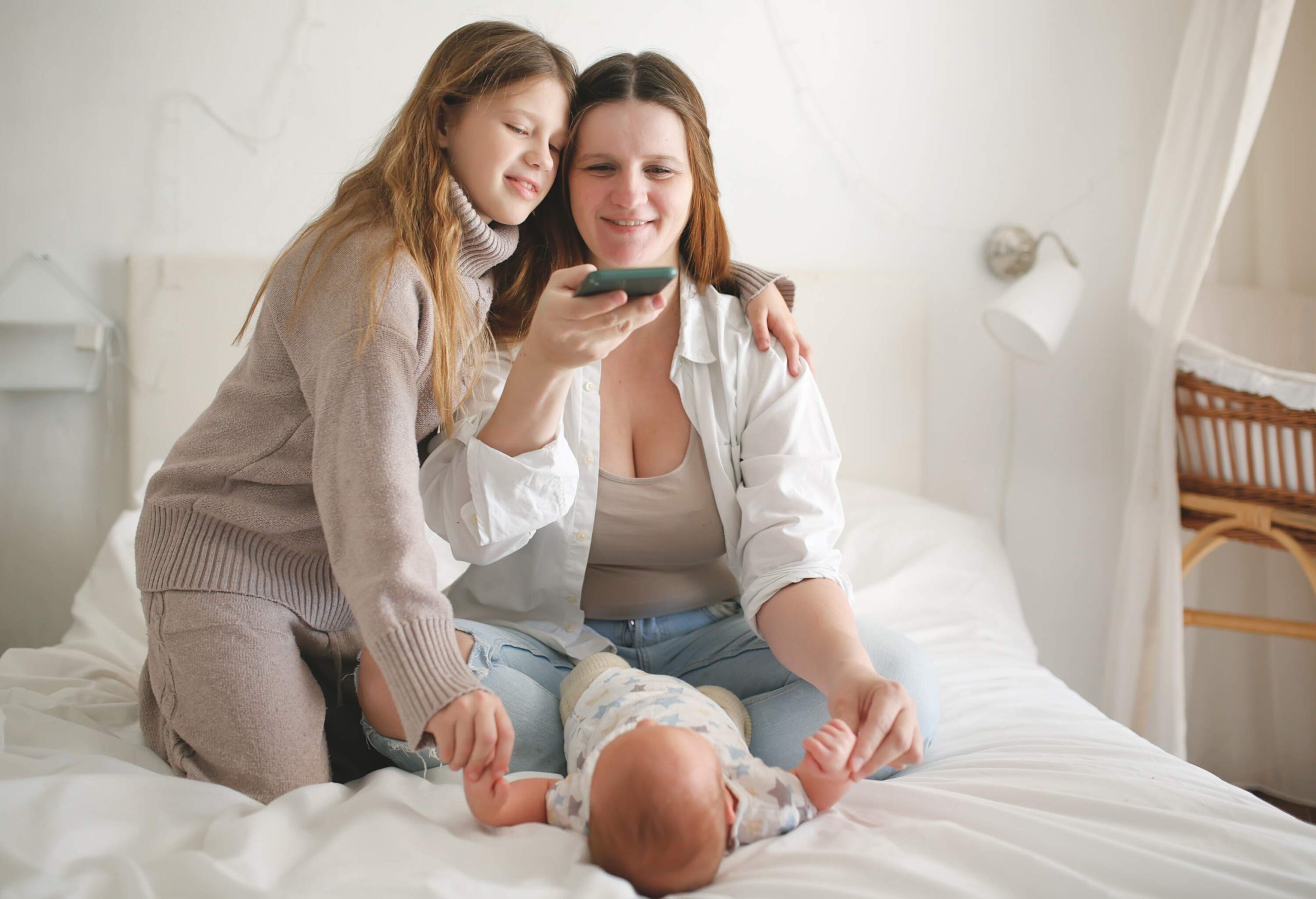 Mother and older daughter take pictures on phone of baby on bed in cozy real bedroom with wicker cradle