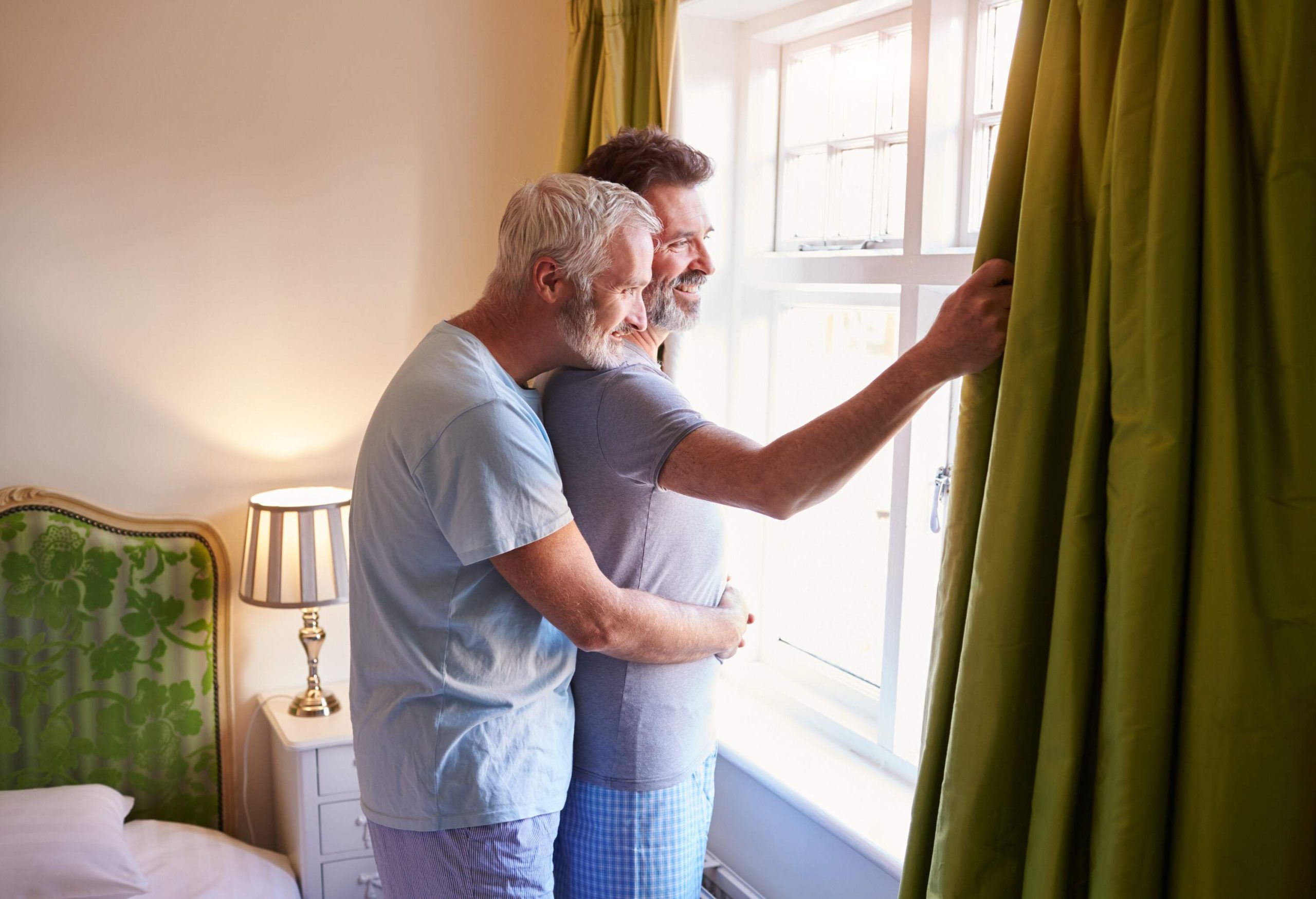 An elderly man gives his lover a back hug while looking out the hotel window.