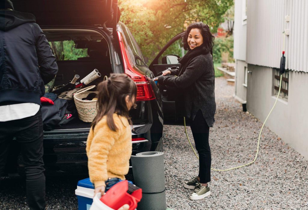 A woman charging an electric car while man and little girl load stuff into the trunk.