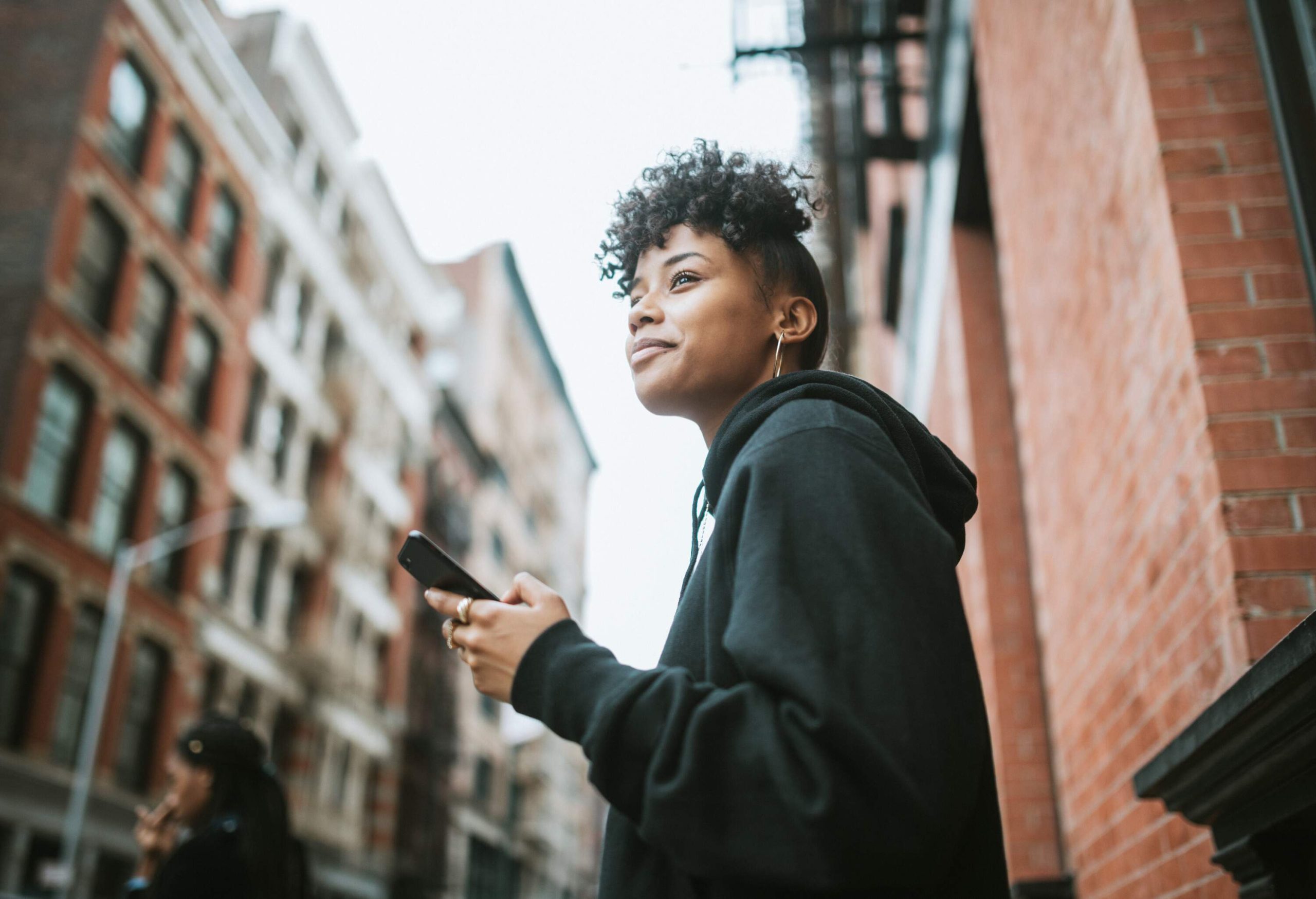 A curly-haired woman in a black hoodie holds her phone as she stands on a sidewalk.