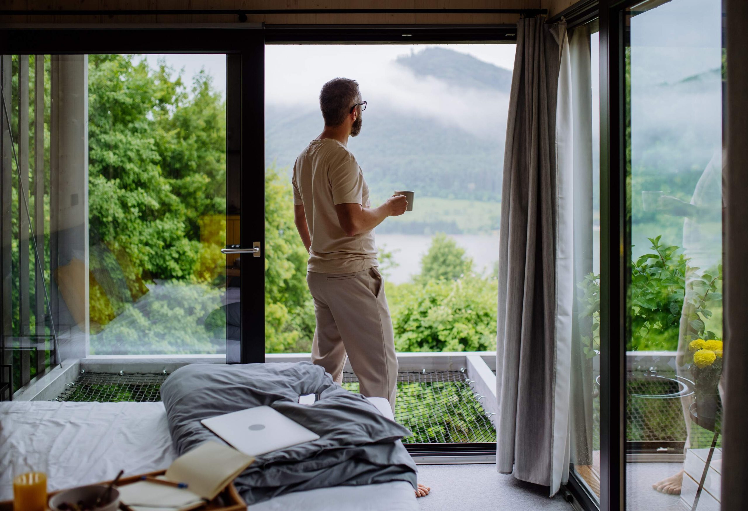 A man holds a cup of coffee as he stands beside a big glass window with vast nature views.