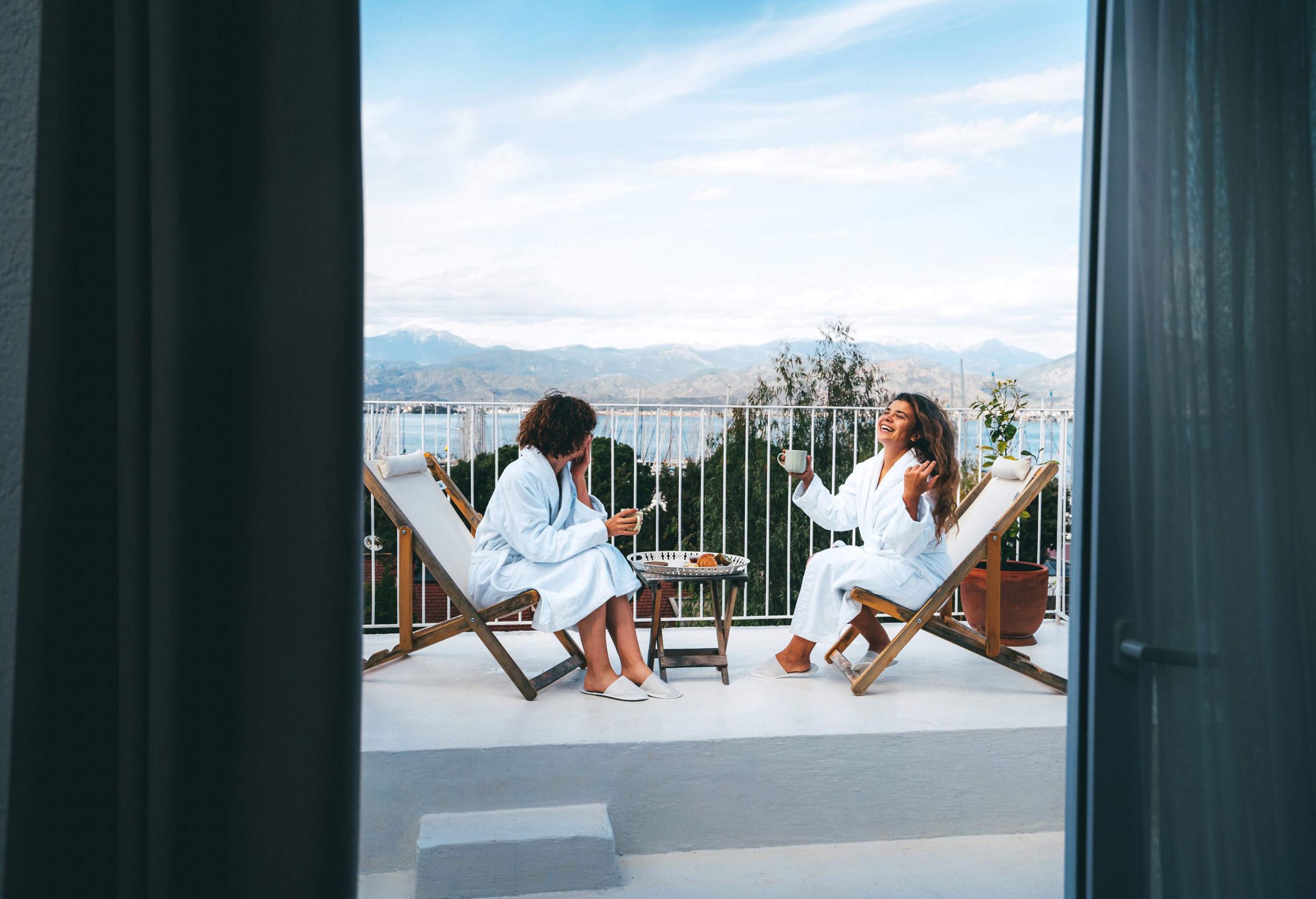 Two friends in SPA bathrobes having a good time drinking tea with the scenic view on the hotel room balcony. Bachelorette party, happiness when spending time together.