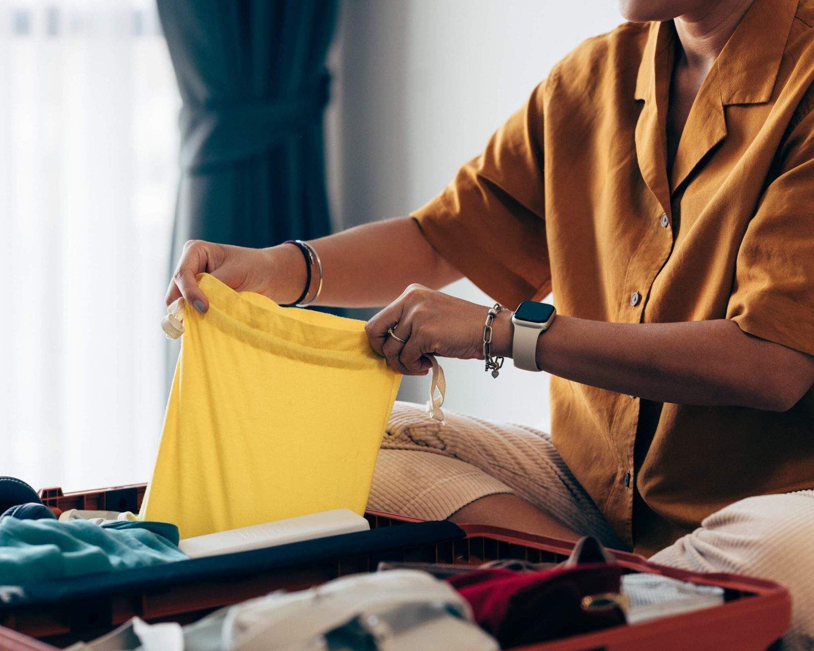 Anonymous woman sitting on the bed in a hotel room, packing her clothes in a suitcase.