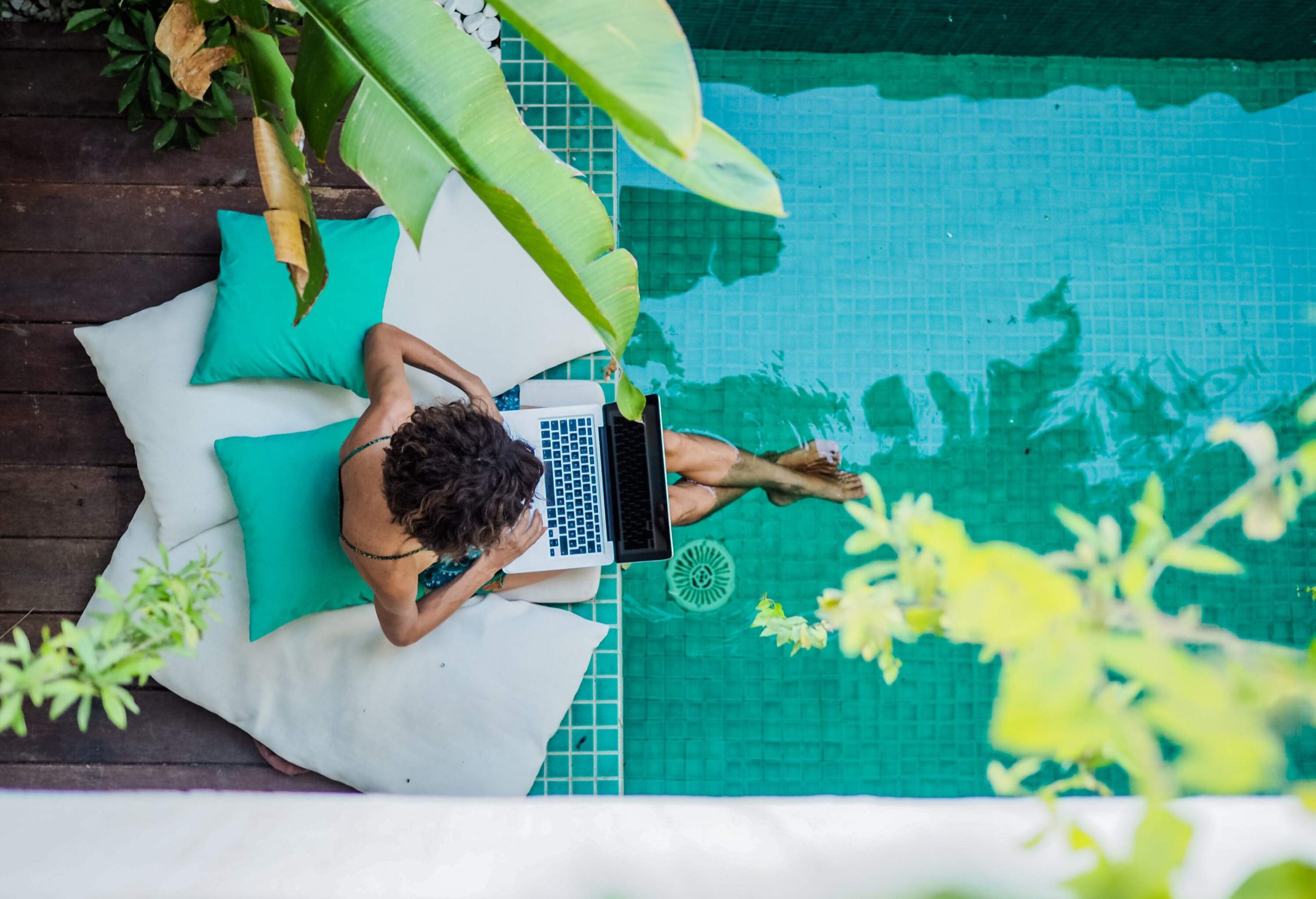 A woman sitting on large cushions on the side of a pool, her feet dangling into the water, working on her laptop.