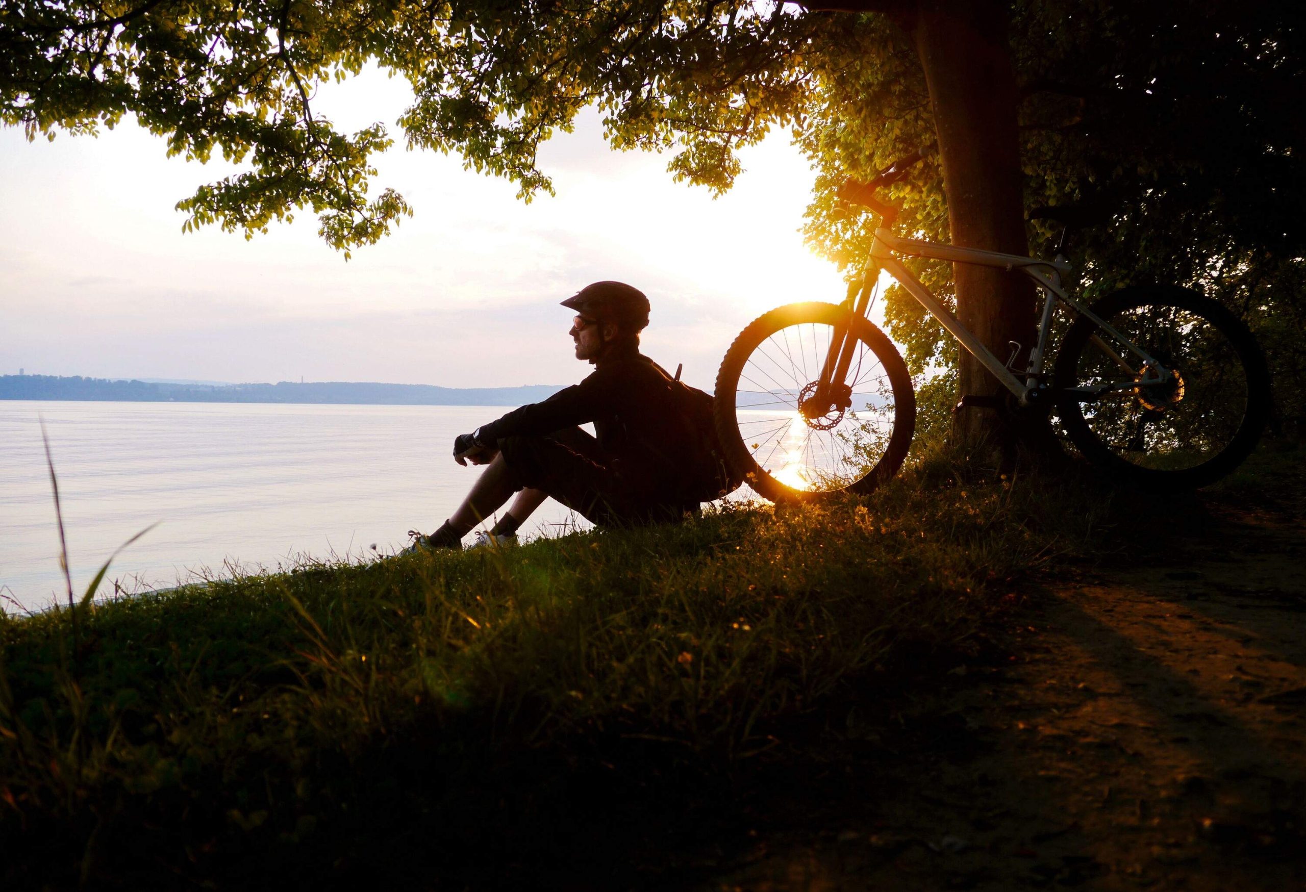 A cyclist sitting on the grass by a lake with his bike parked against a tree.
