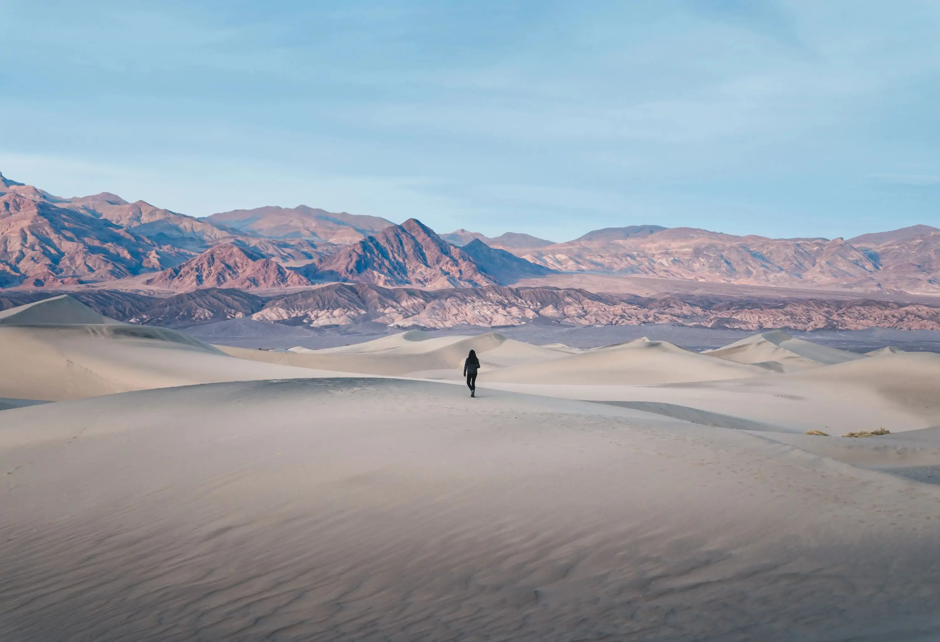 A person walks across a vast sand dune in a desert with views of rugged mountain range.