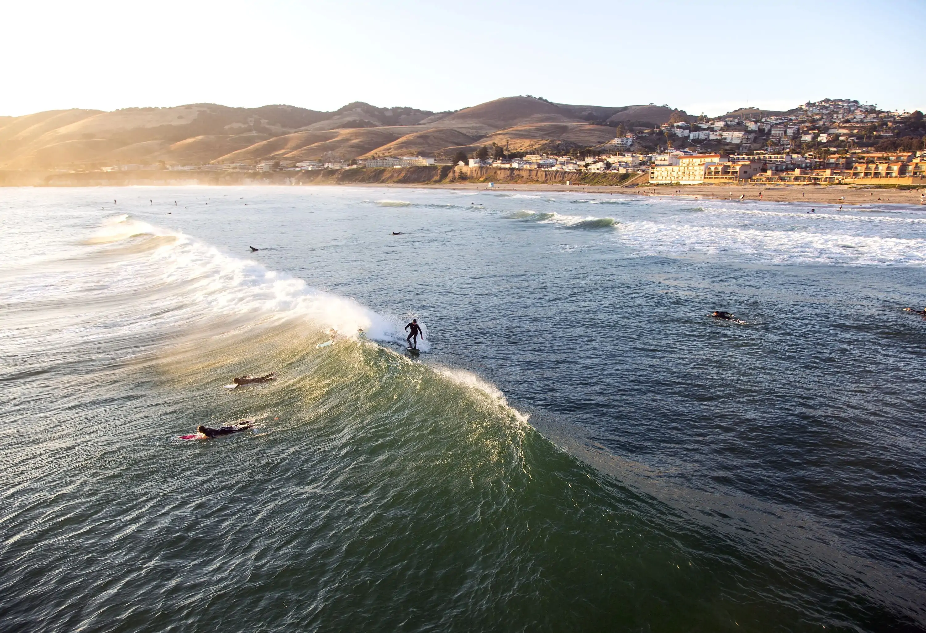 A surfer rides a raging wave on a beach with distant views of a coastal village.