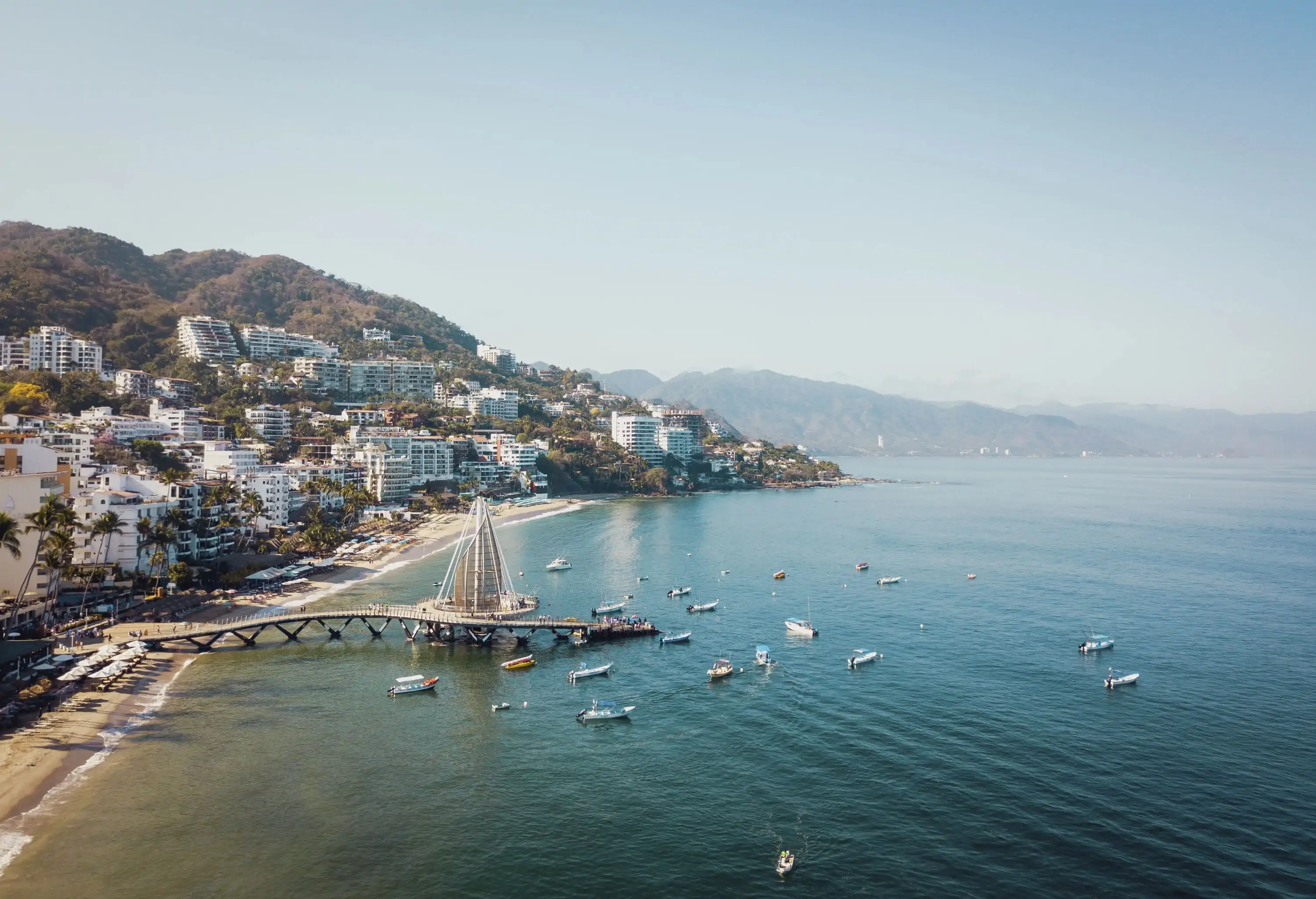 A picturesque coastal town boasts a bustling pier with vividly coloured boats, while buildings cascade down the hillside, embraced by verdant surroundings.