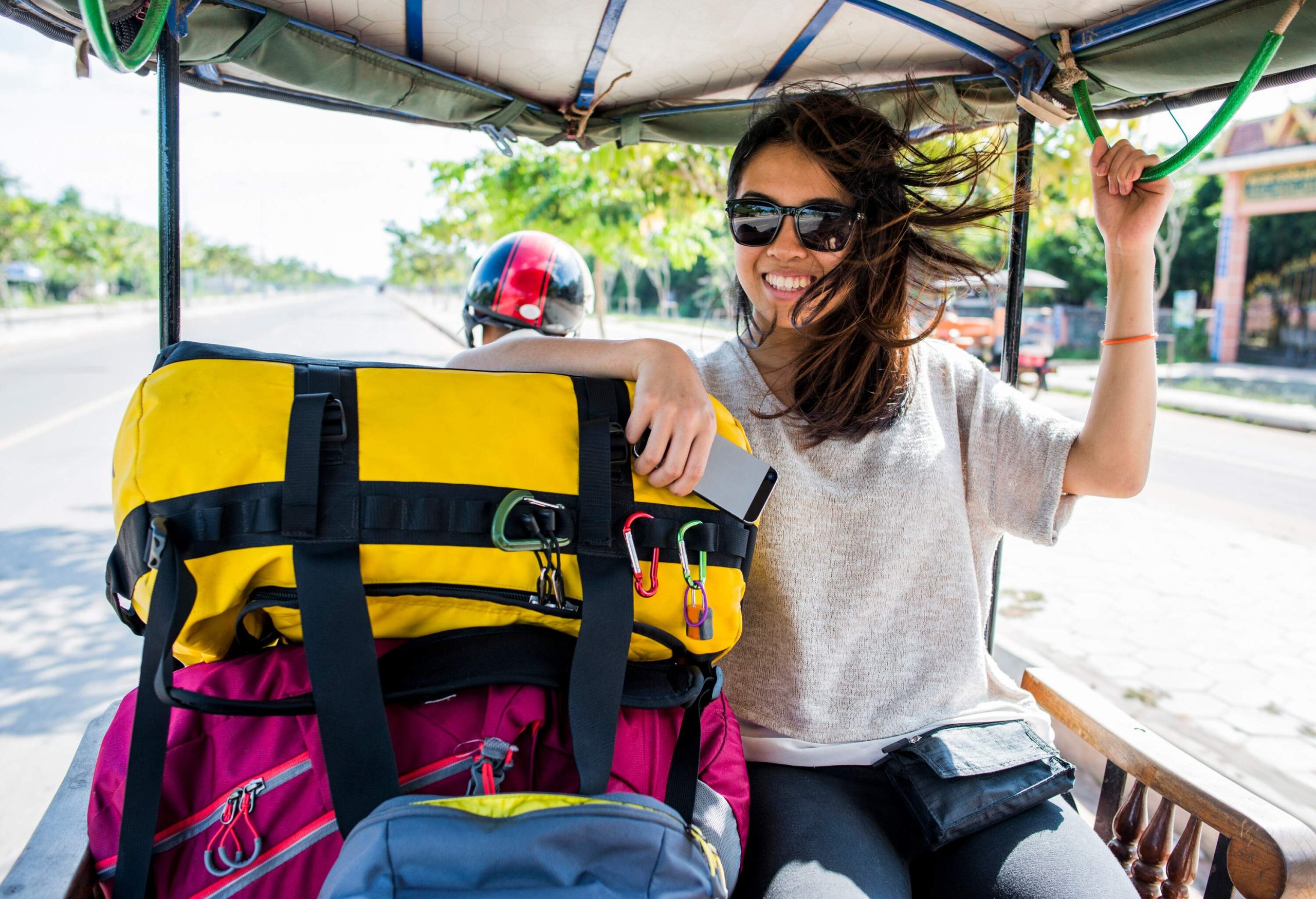 A woman in sunglasses and casual clothes smiles as she rides at the back of a vehicle with a pile of luggage.
