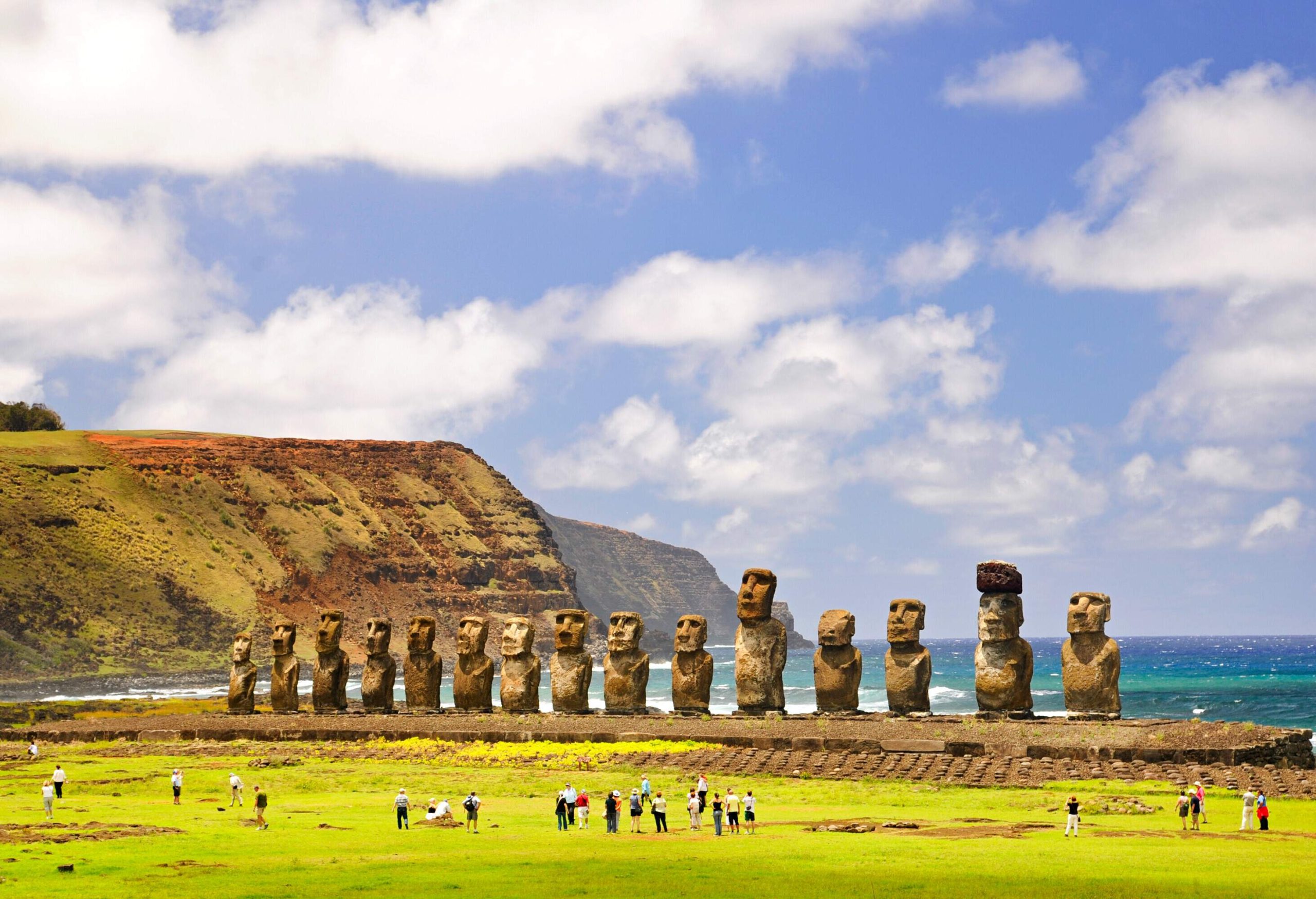 The iconic 15 Moai statues, large stone monoliths carved by the Rapa Nui people, stand tall along the coastline, with visitors exploring the nearby turf and a dramatic coastal cliff serving as the backdrop.