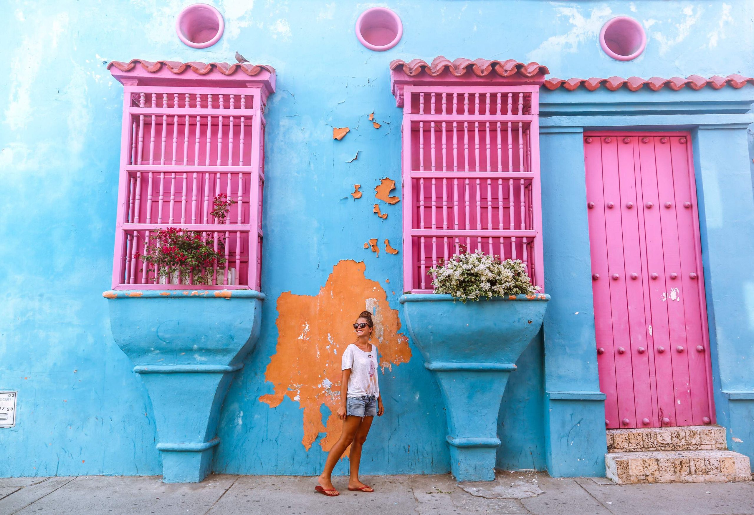 A person wearing sunglasses strikes a pose in front of a light blue building with enclosed windows and a wooden door on the side.