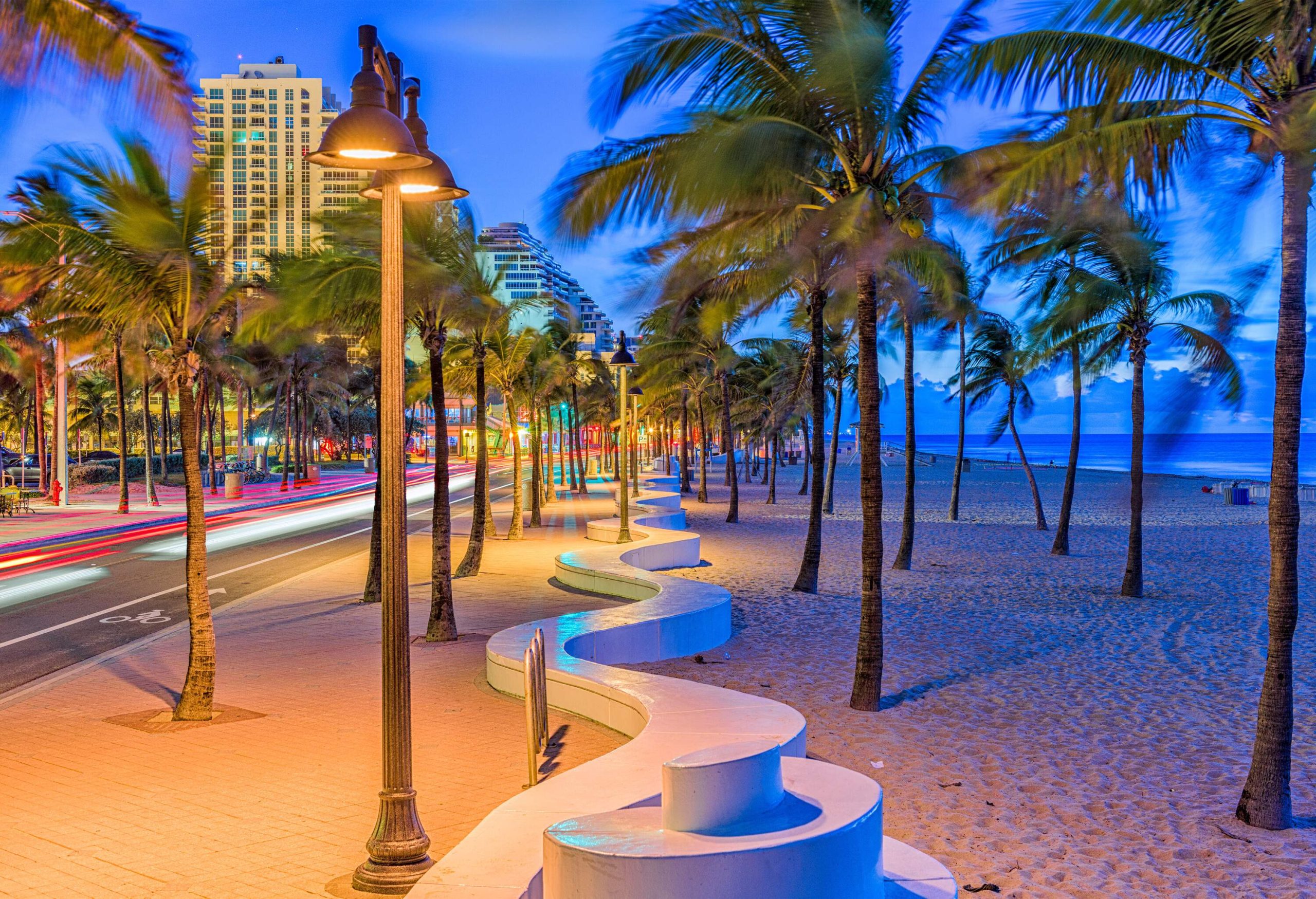 A long winding concrete white bench concealed under the palm trees and lampposts lining the seaside road against the scenic blue sky.