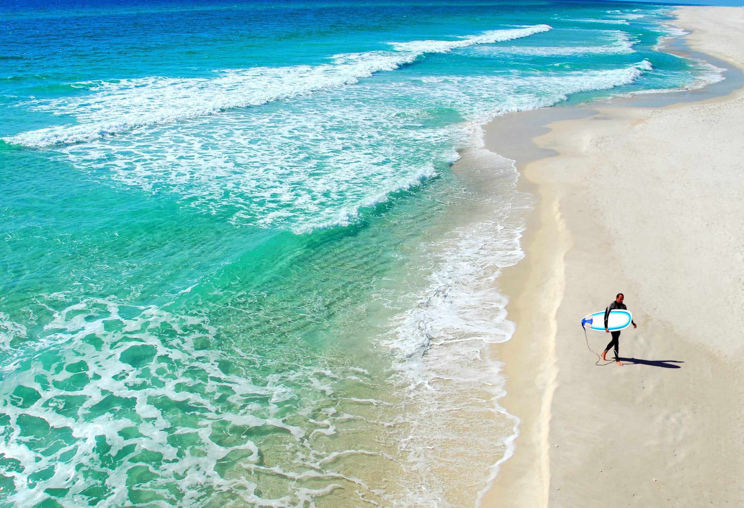 A man with a surfboard walks on the shore of a wavy beach.