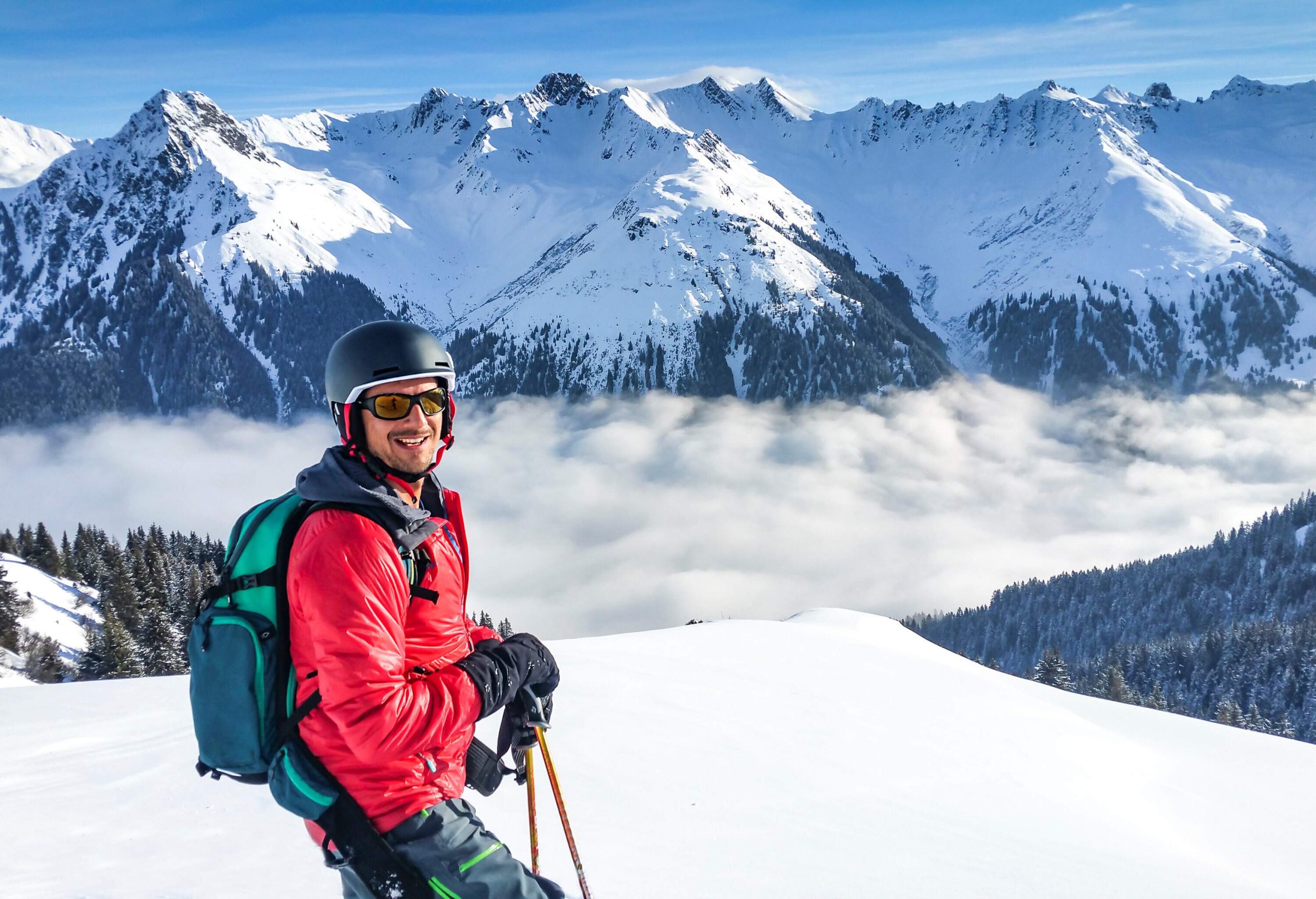 A smiling skier with a red jacket and helmet stands on a snowy hilltop against a background of thick fog at the foot of snow-covered mountains.