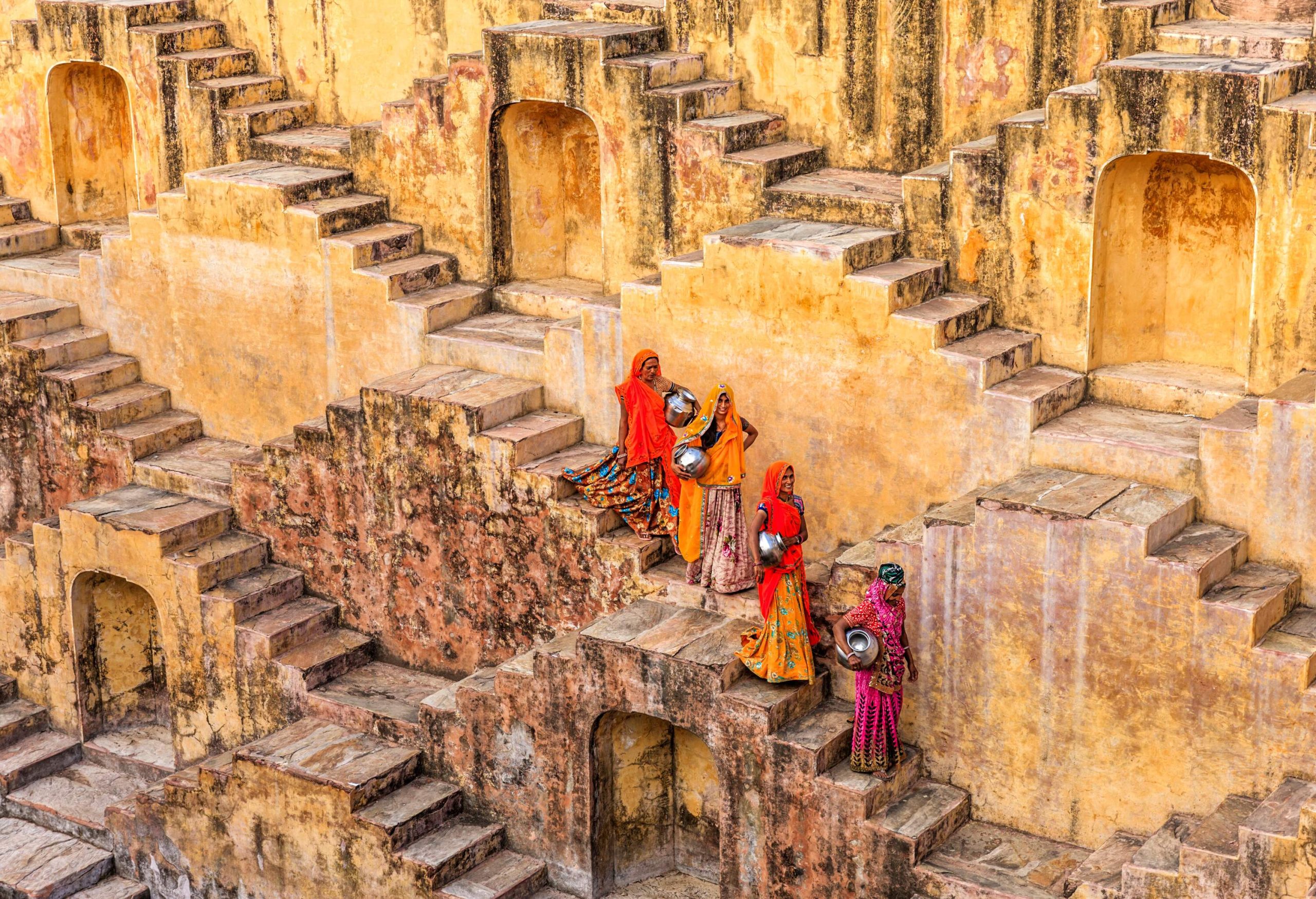 Indian women gracefully carry water from a stepwell, navigating a series of stairs, while the faded paint of the surroundings adds a touch of rustic charm.