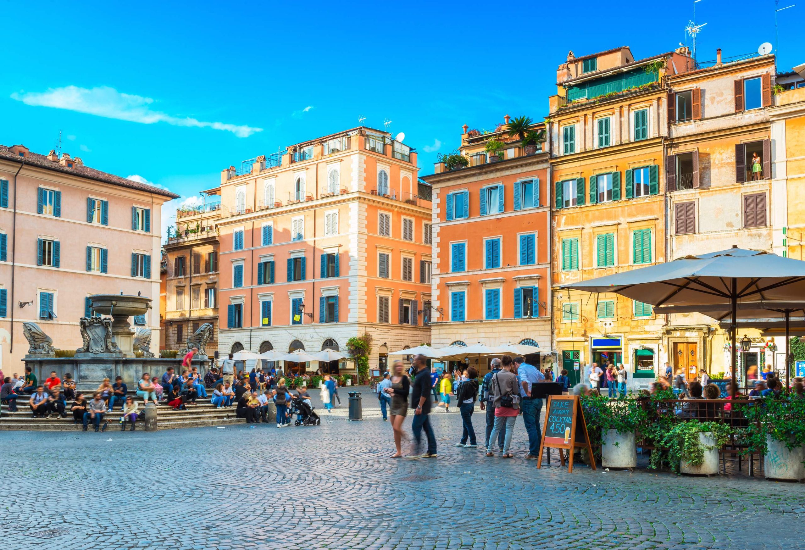 A bustling public square with people sitting on steps around a fountain and strolling past outdoor cafes.