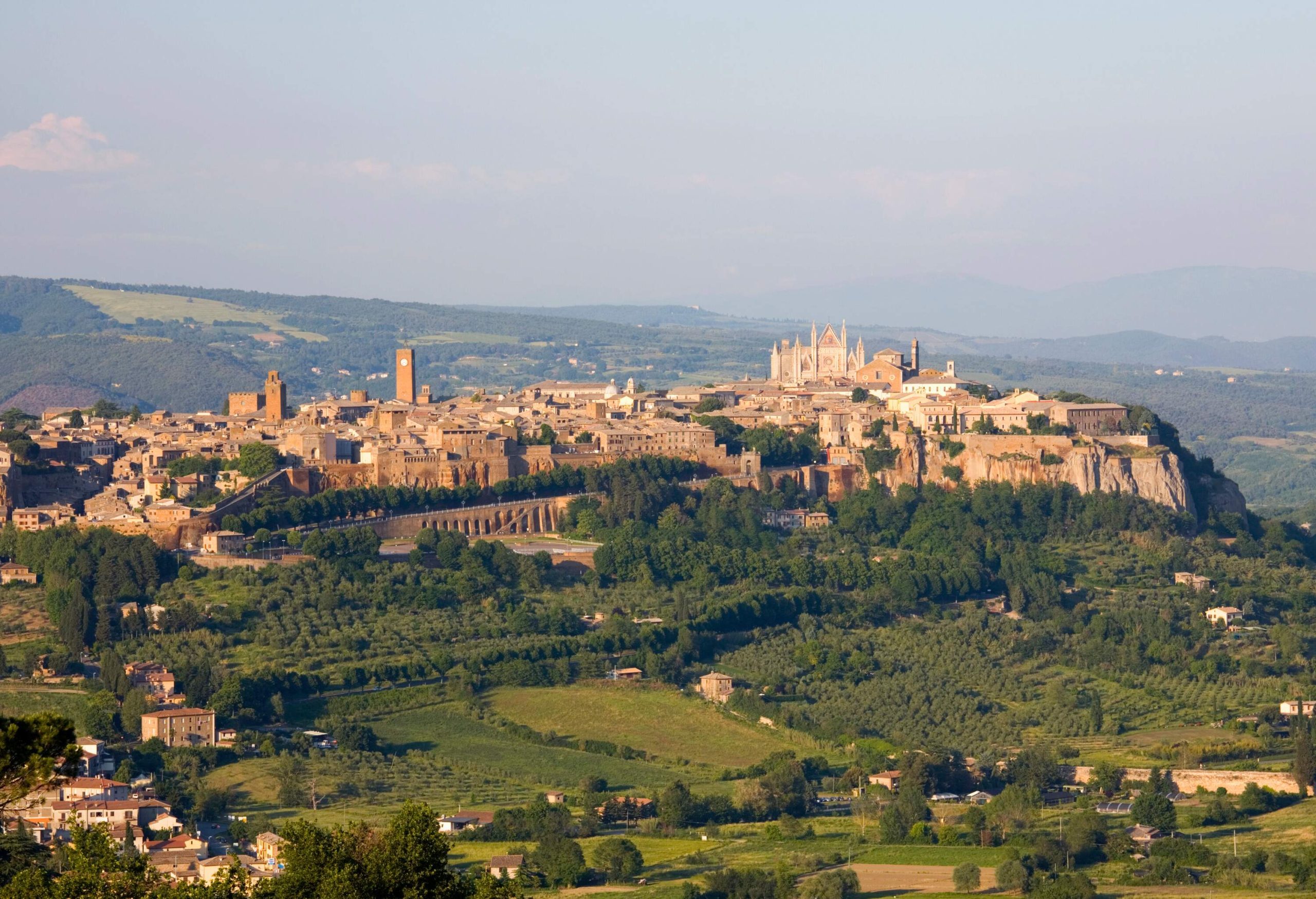 A landscape of verdant land and an old city with classic buildings, houses and churches on a rock cliff.