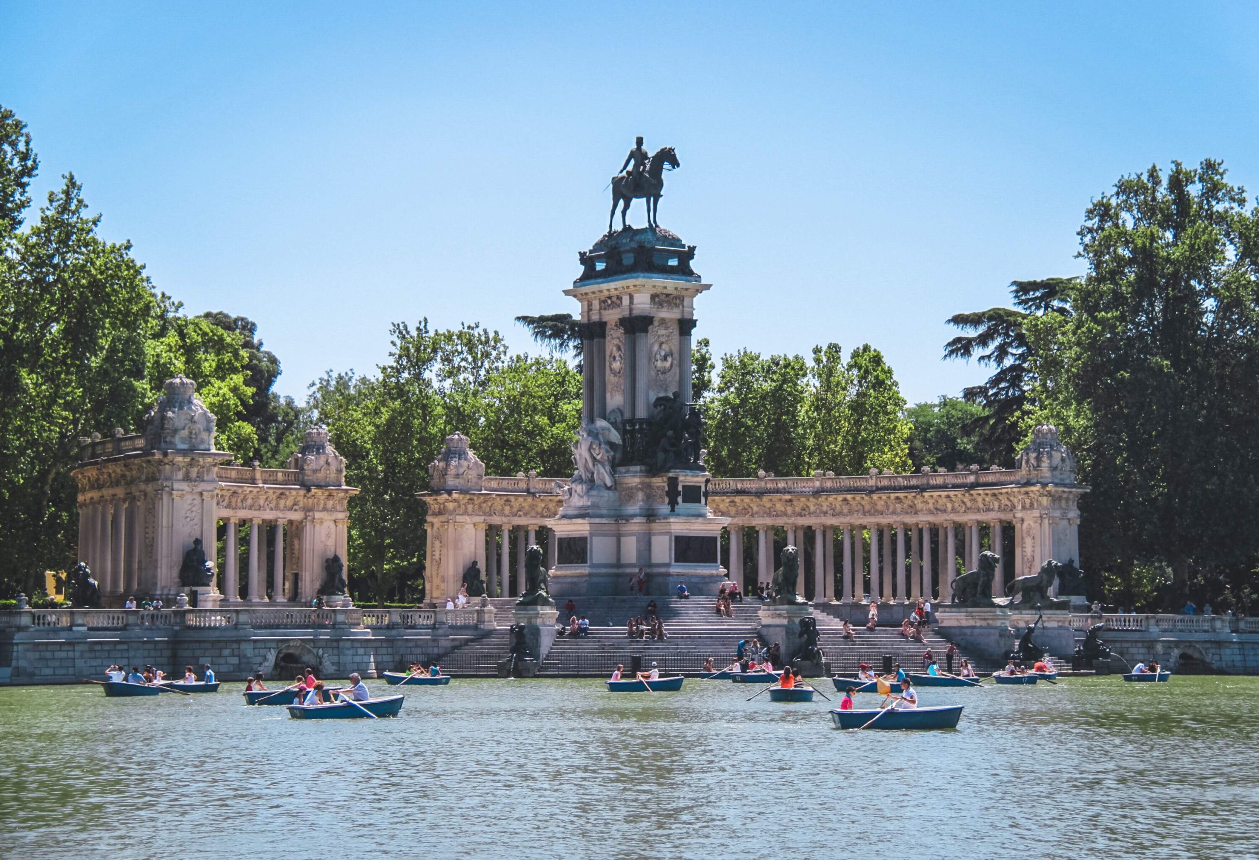 People on small blue boats paddle on the artificial lake in front of the monument of the equestrian sculpture of a king.