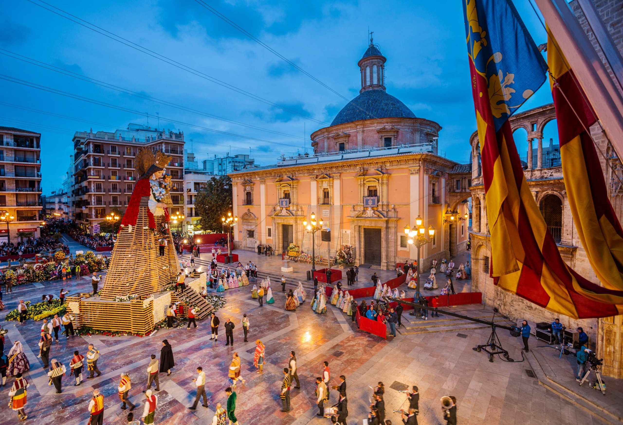 A festival that takes place on a plaza, with a huge wooden figure of the Virgin Mary in the middle wearing a red mantle, people standing around it giving it flowers, and some wearing traditional attire.