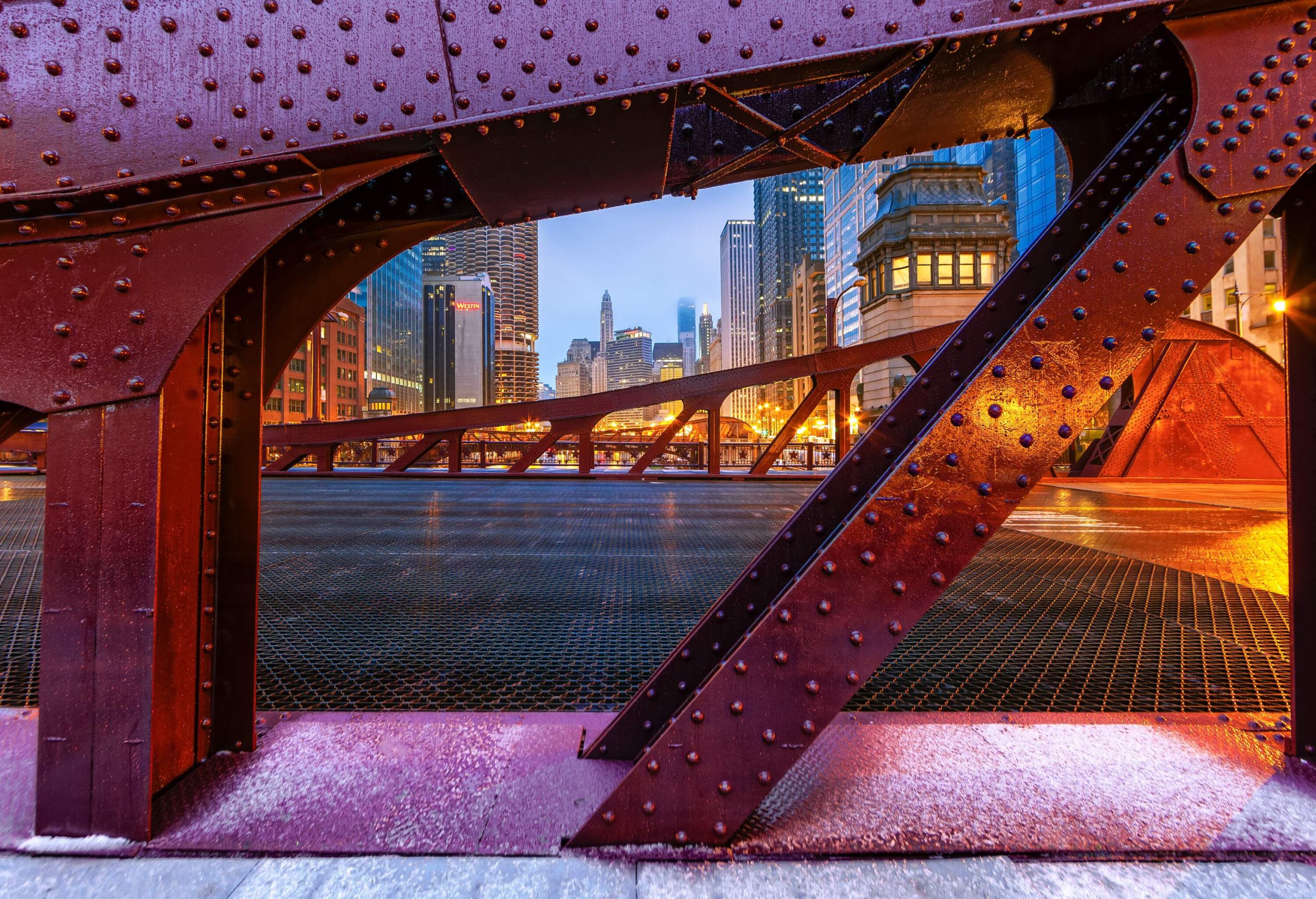 Massive metal foundations of a bridge with multiple studs and views of an illuminated cityscape.