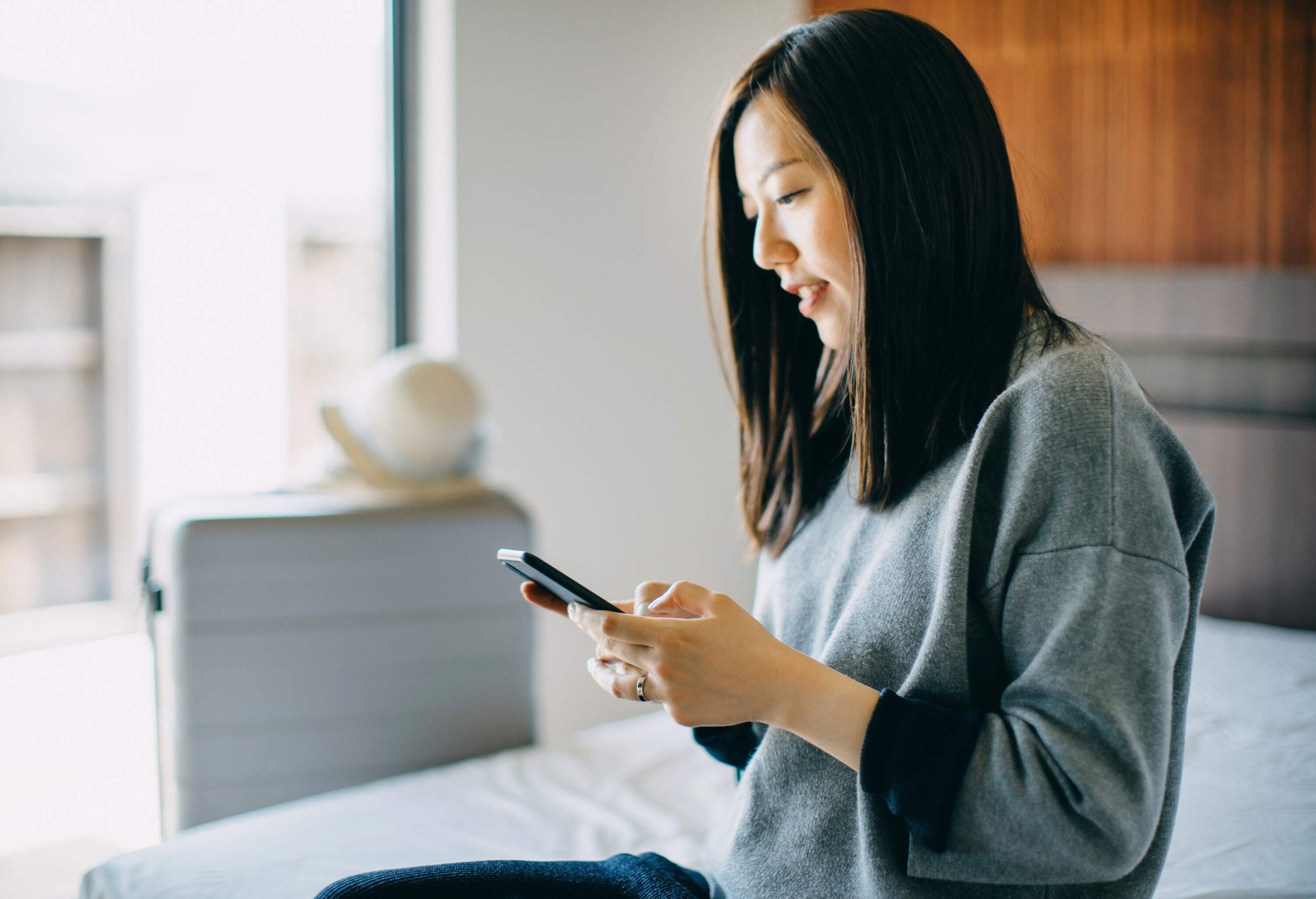 A dark-haired woman in a grey shirt sits on the bed as she uses her phone.