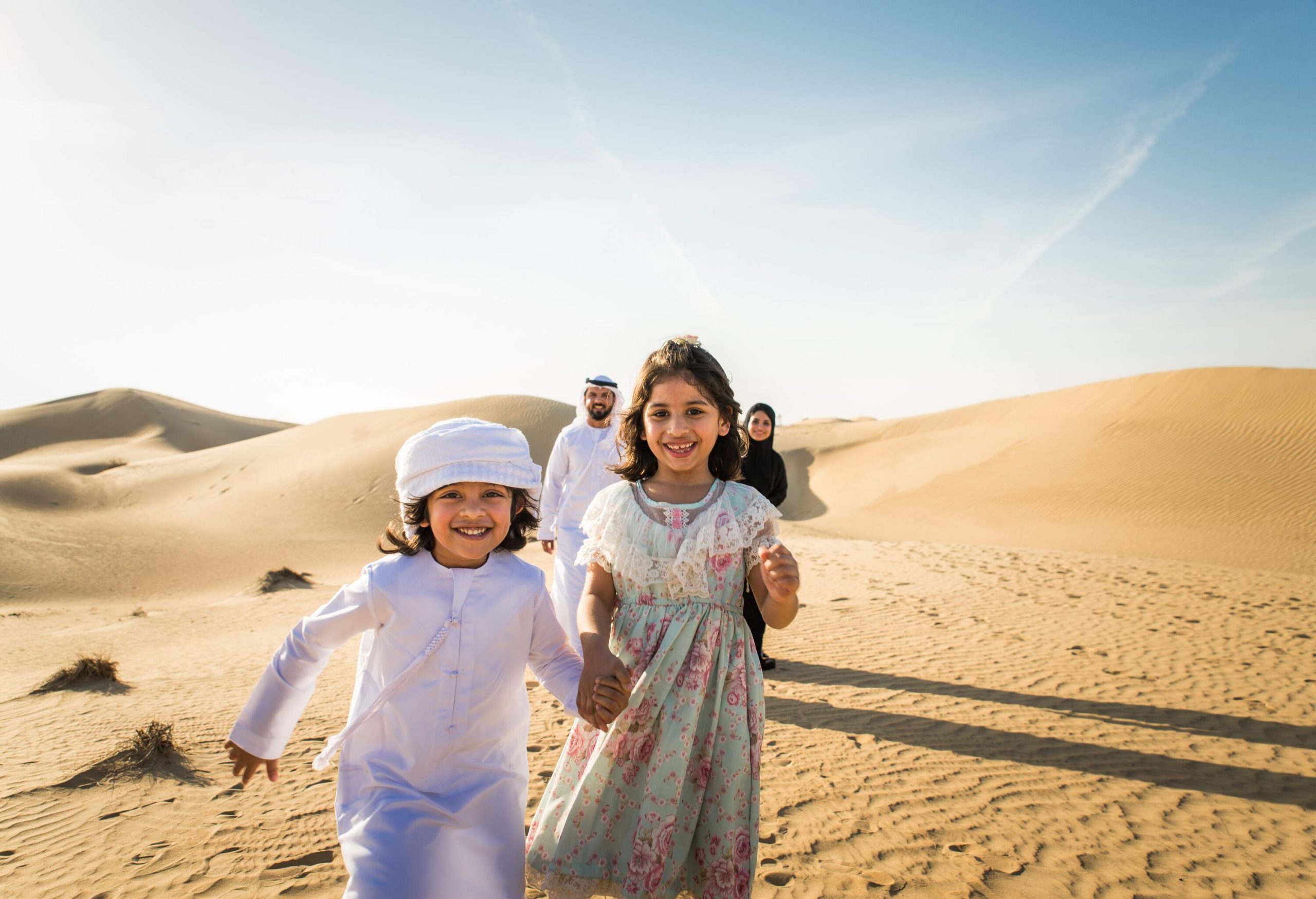 Arab family of four in the desert, the mother, father and son are dressed in traditional Muslim clothes and the daughter is wearing a colourful dress.