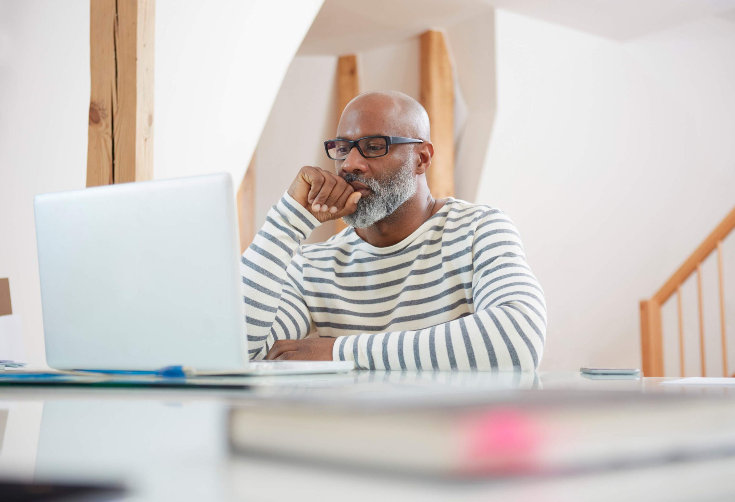A bald man in glasses and striped sweater watches something on a white laptop.