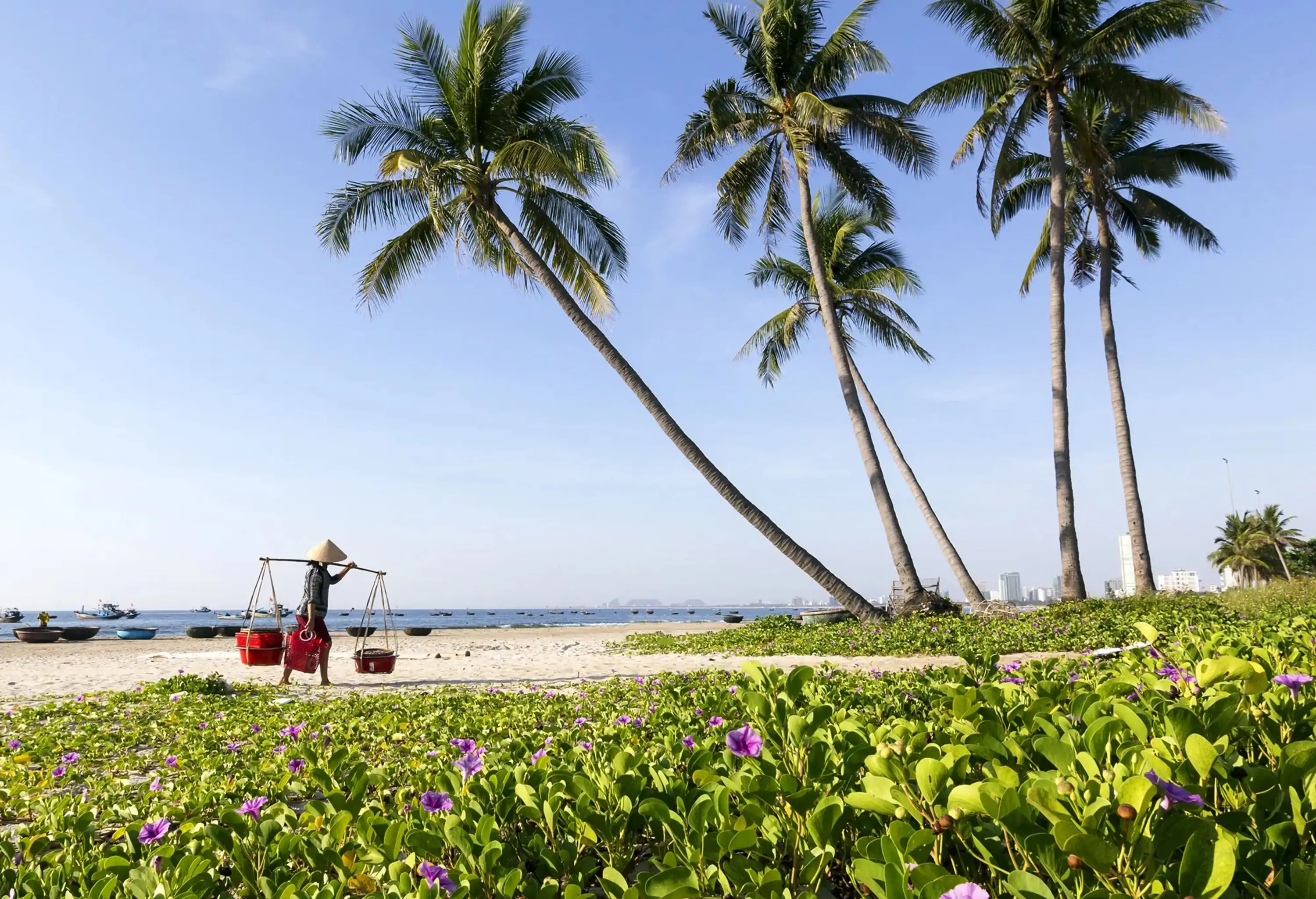 A man carrying baskets with a shoulder pole as he walks toward a beach covered in creeping vines next to a grove of palm trees.