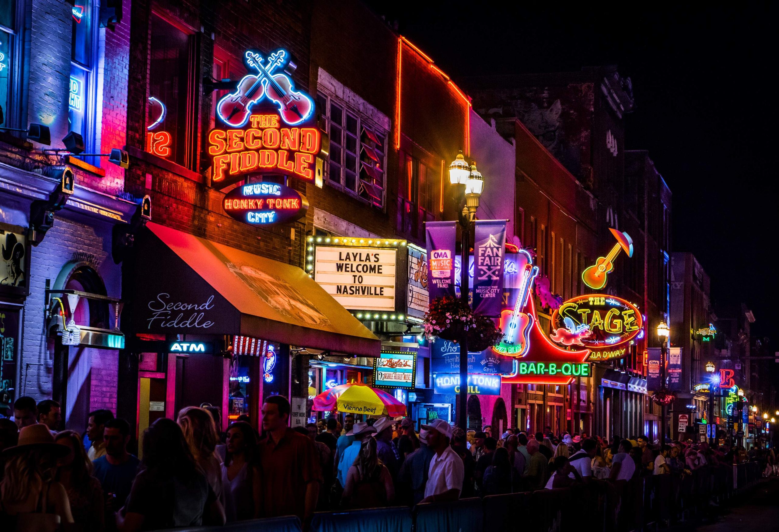 The lively sidewalk of the entertainment district teems with a vibrant crowd, as the mesmerising glow of neon lights from overhead signage adds an electric atmosphere to the bustling scene.