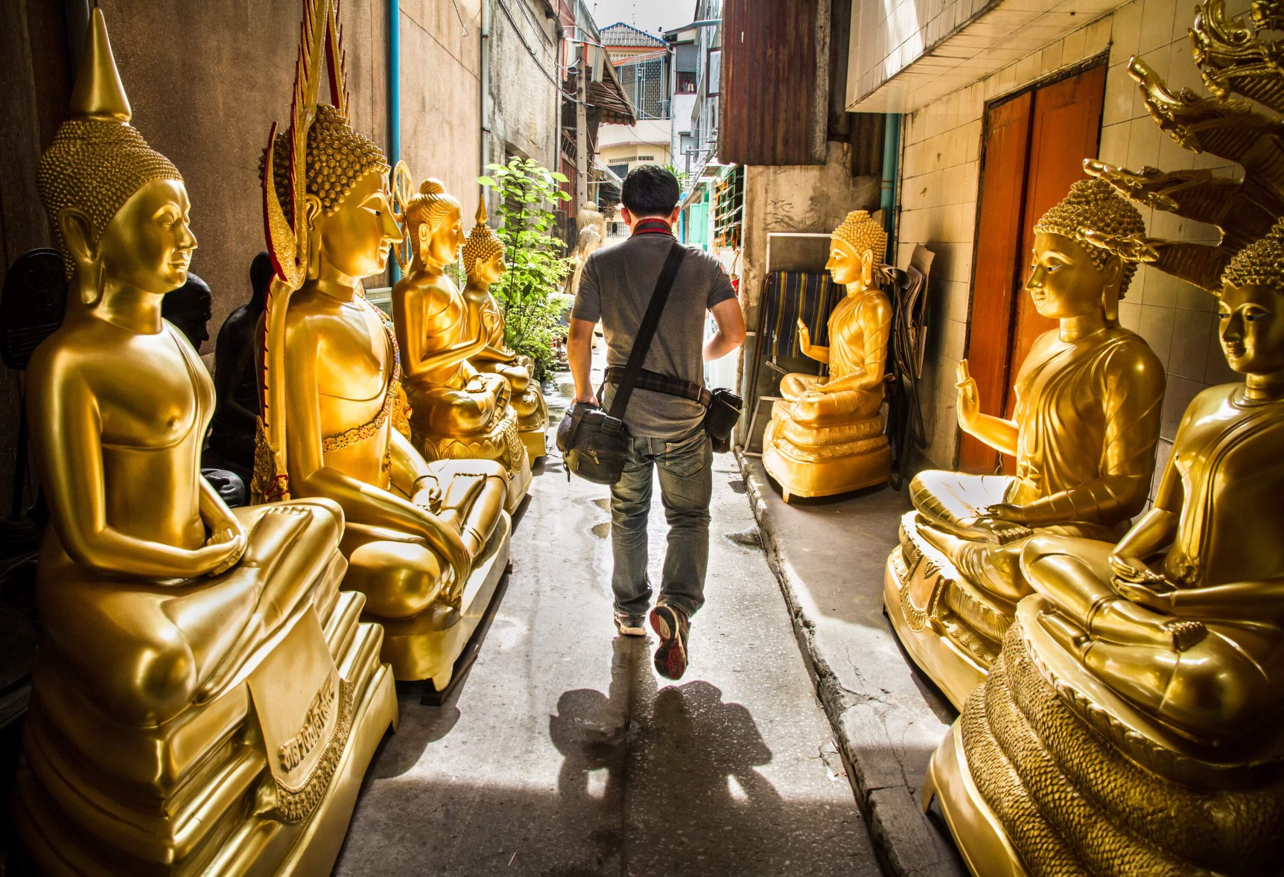 A man carrying a camera walks on a narrow street with golden Buddha statues flanked on both sides.