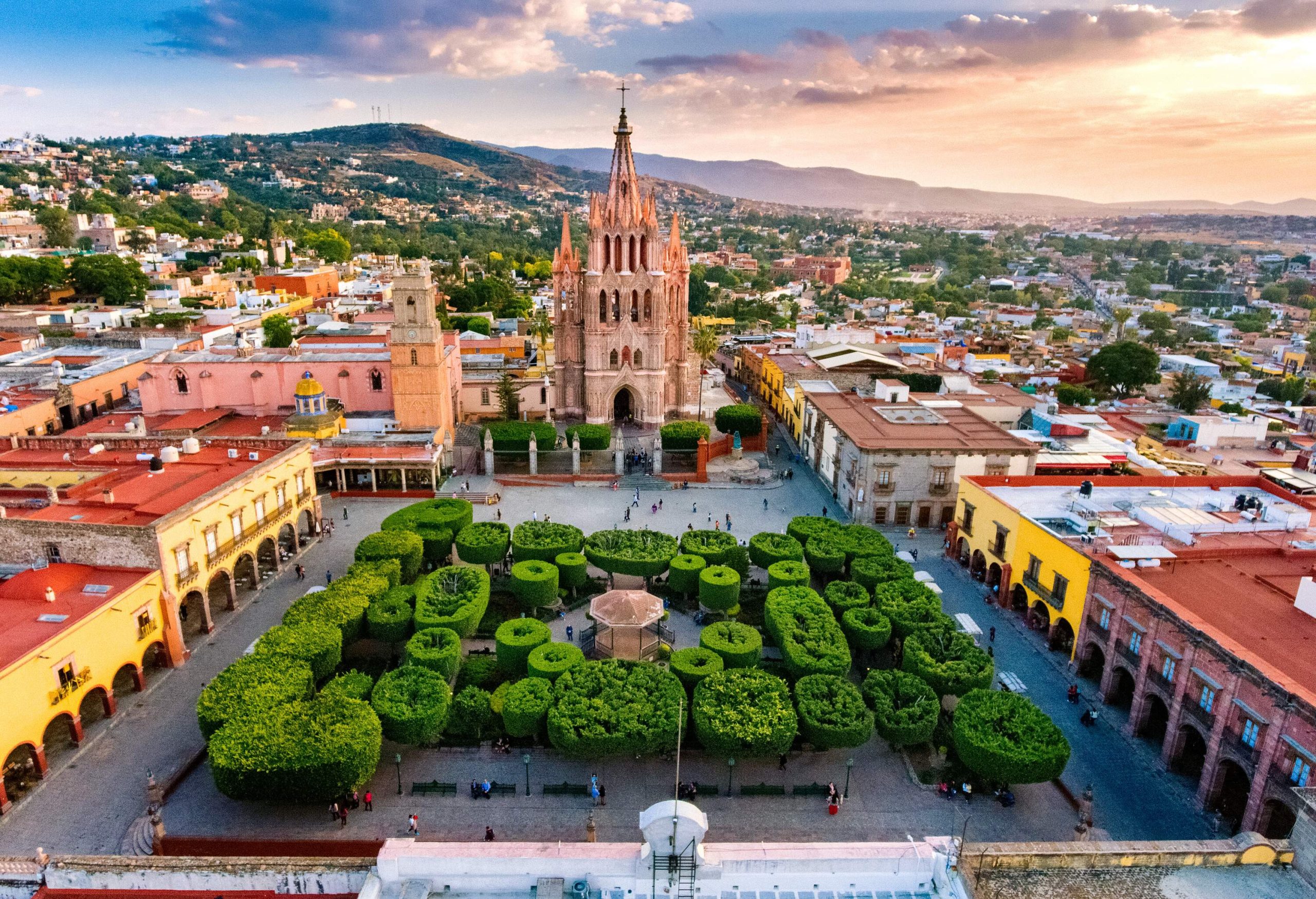 A lush garden of neatly trimmed trees in the centre of a city square surrounded by classical buildings and a neo-Gothic 17th-century church.