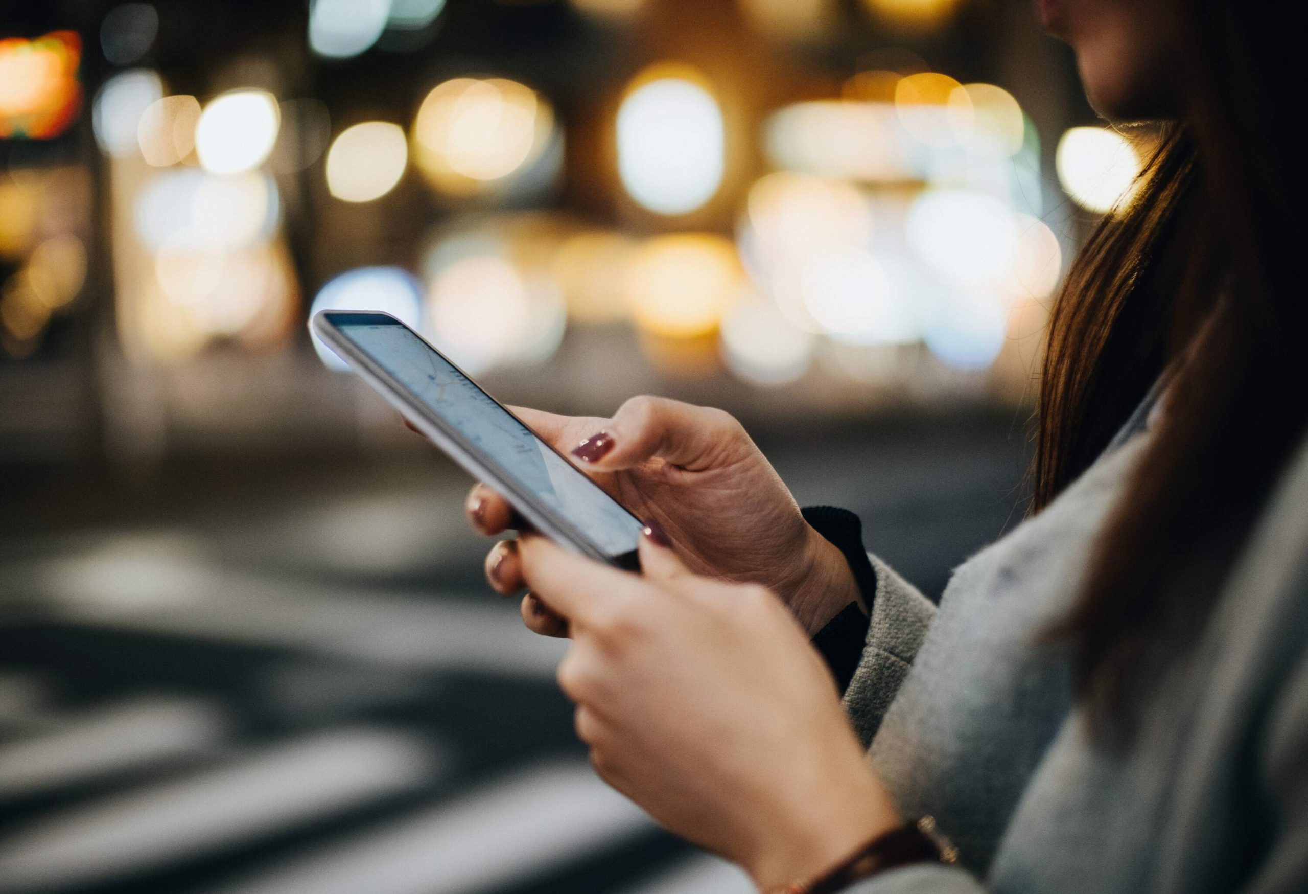 A woman using her smartphone while standing on a busy sidewalk with the blurry dazzling light in the background.