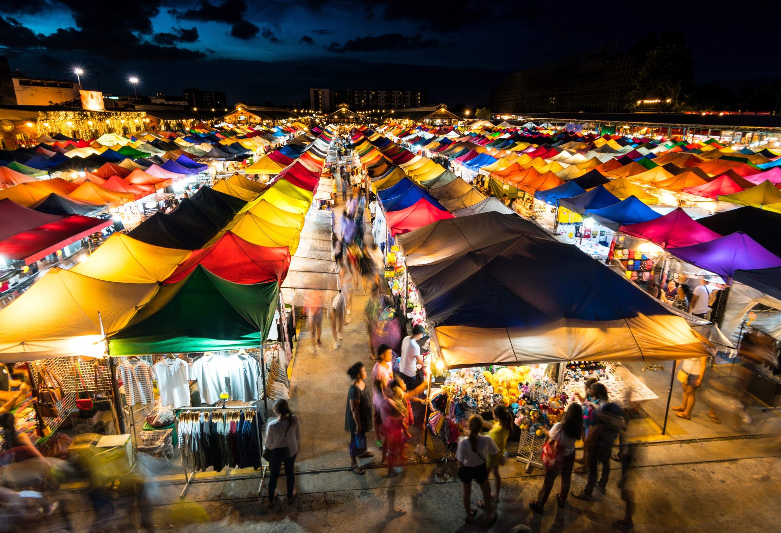 A bustling crowd outside the colourful and brightly illuminated market stalls.