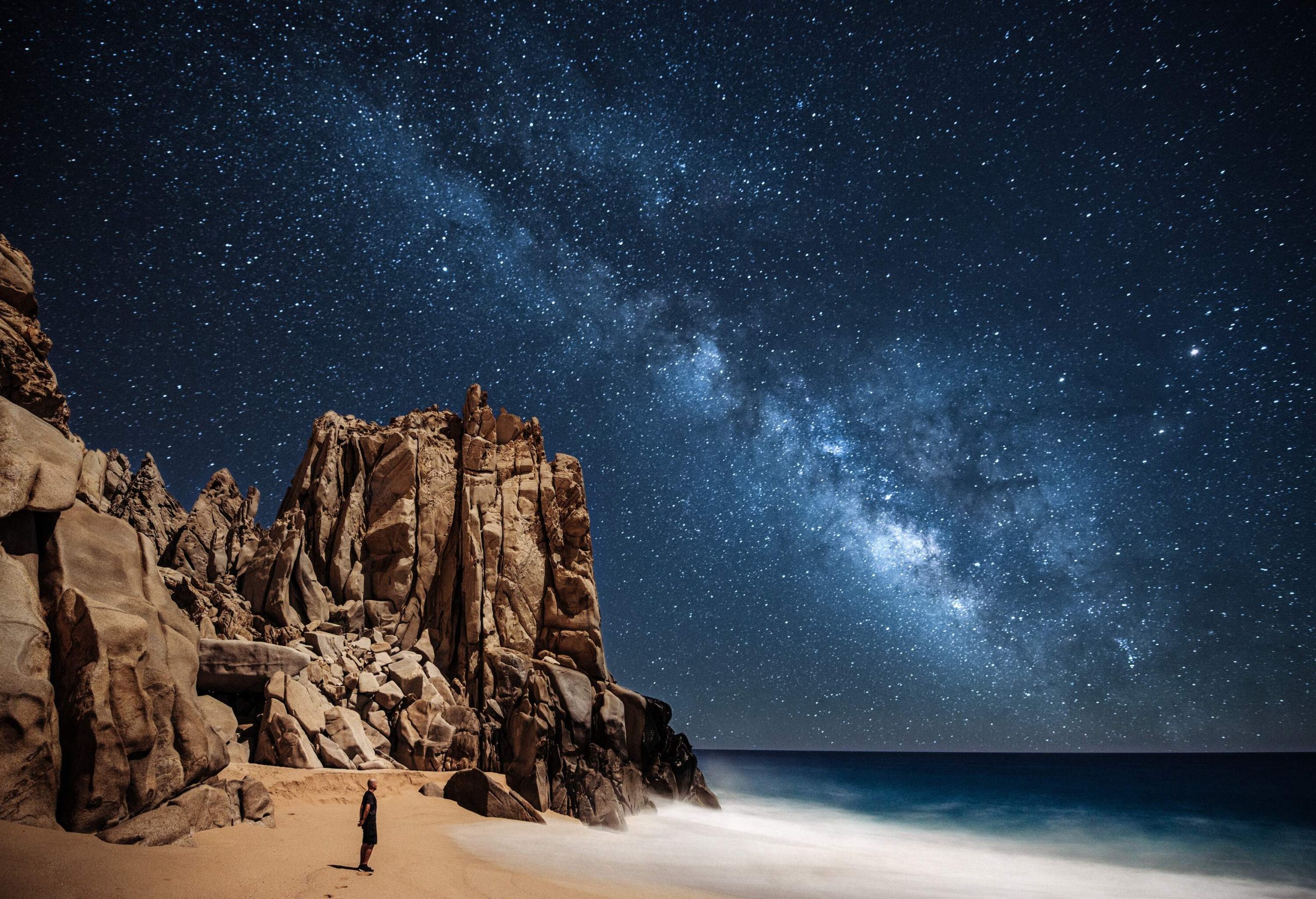 A man gazes at the stars in the calm sky on a scenic beach along steep, rugged rocks.