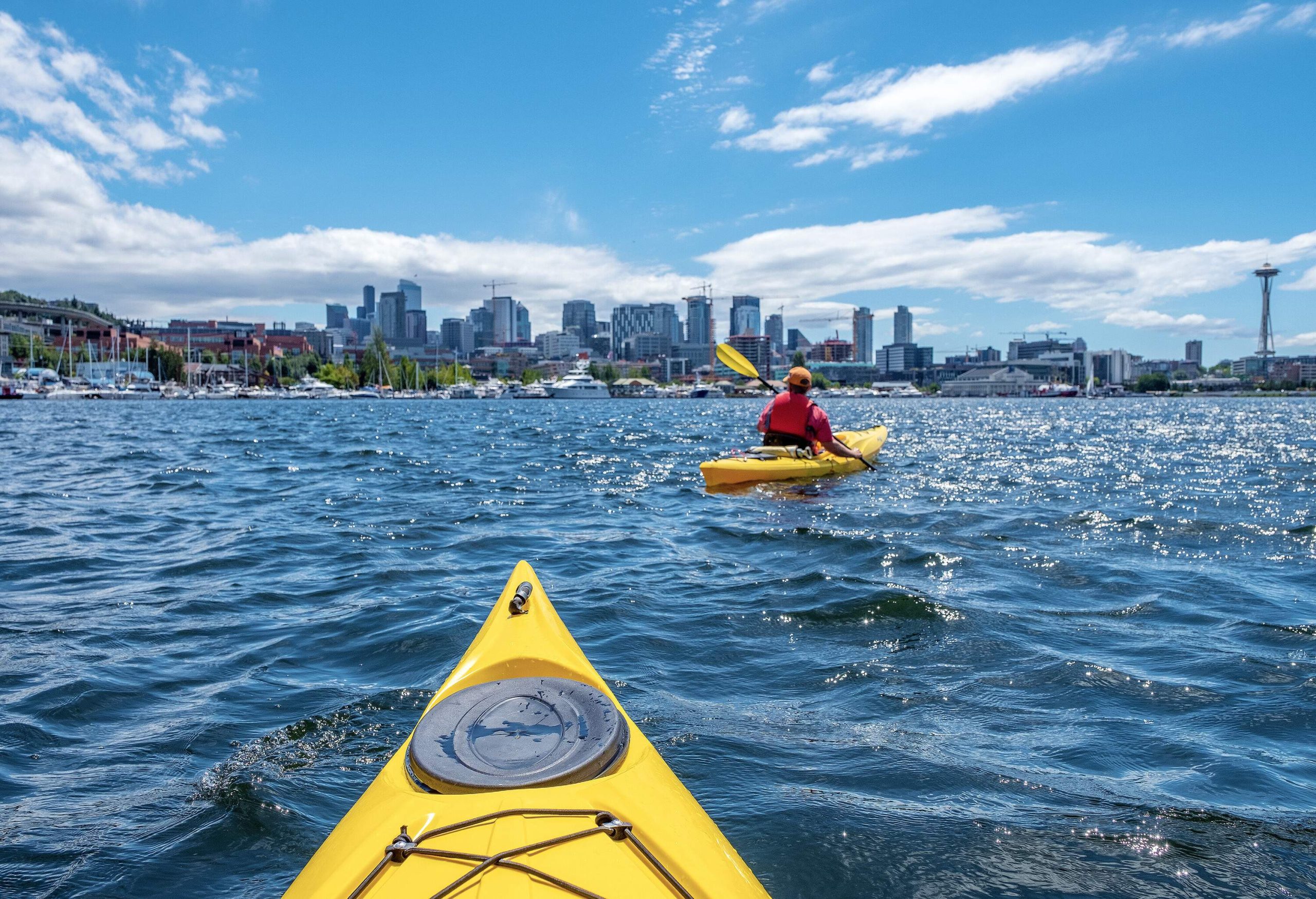 People on yellow kayak boats paddle in the shimmering lake with a view of urban cityscape against the blue sky.