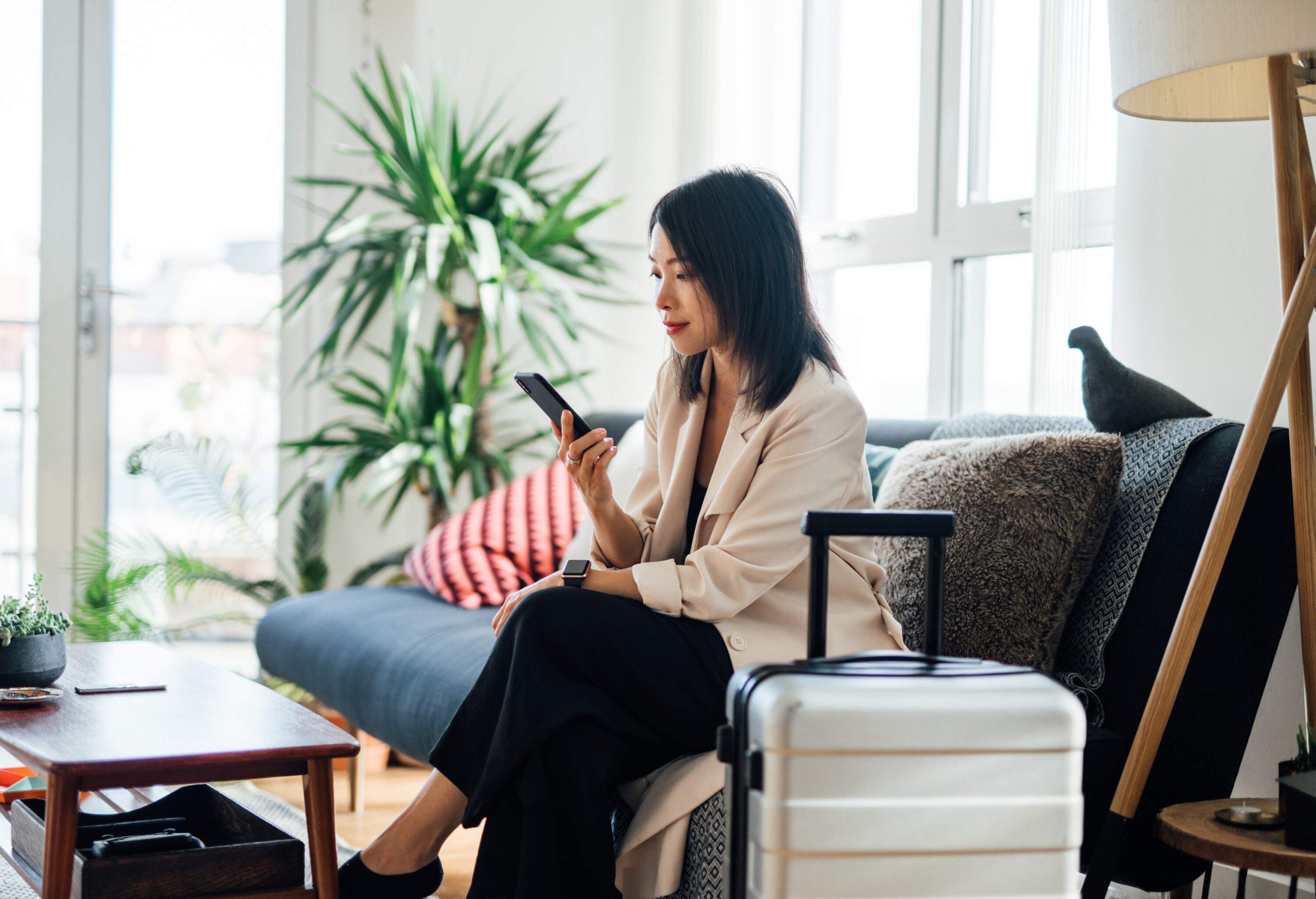 A focused businesswoman sits cross-legged on a sofa with her luggage nearby, engrossed in her mobile phone.