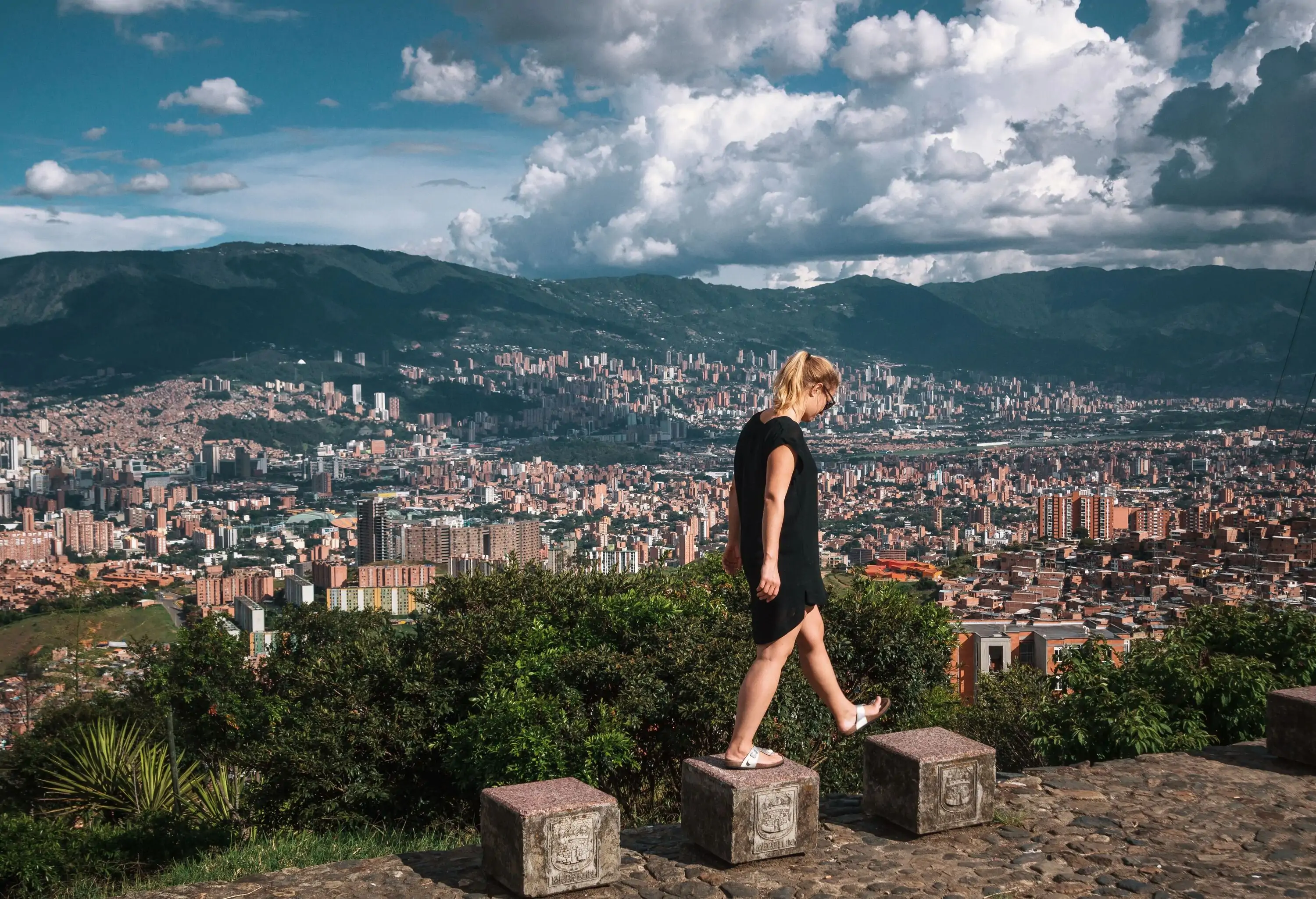 A person in a black dress is joyfully skipping on blocks, overlooking a vast cityscape below and a majestic mountain range in the distance.