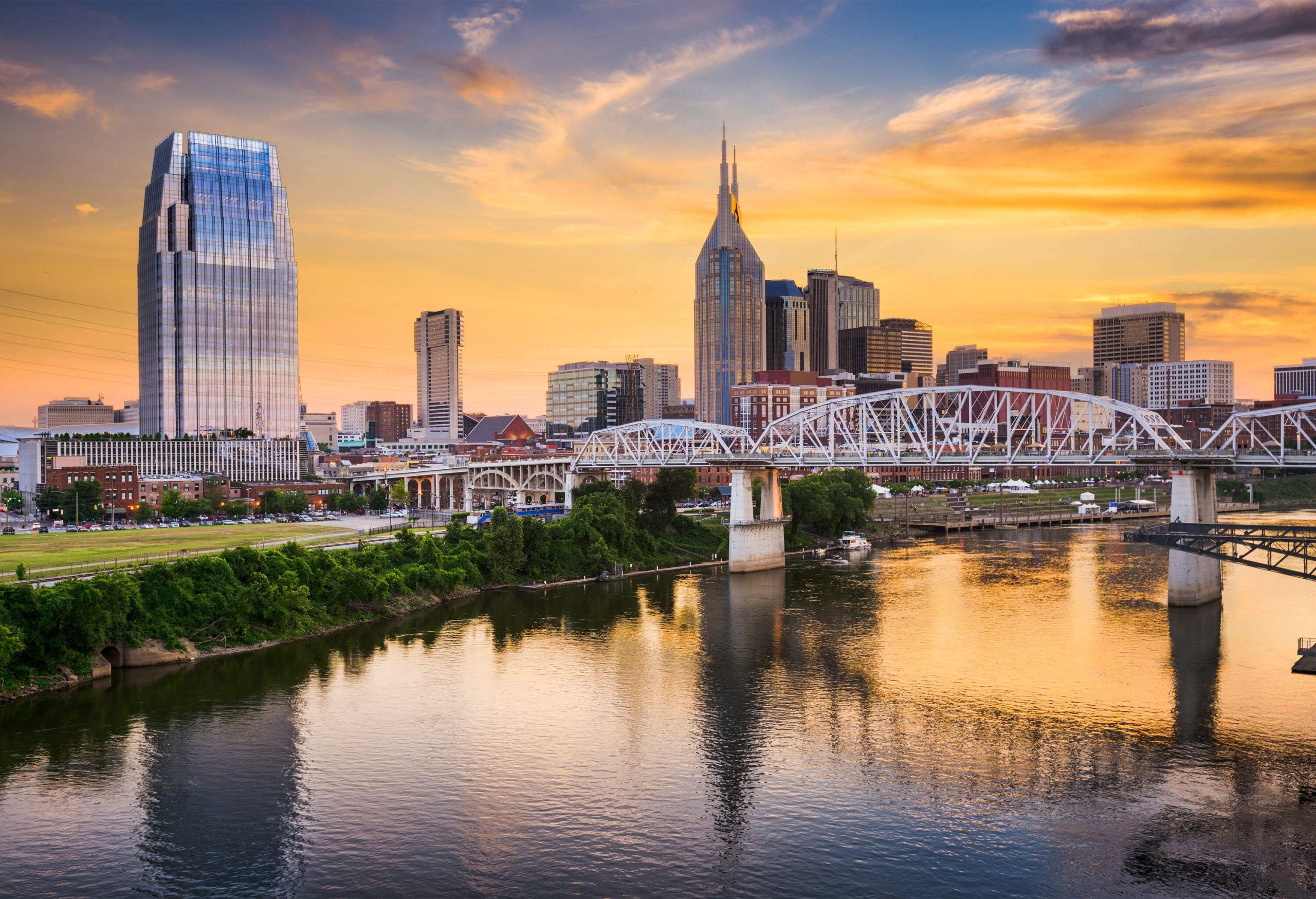 A bridge with arched ribs across a calm river against the backdrop of modern skyscrapers under the orange skies.