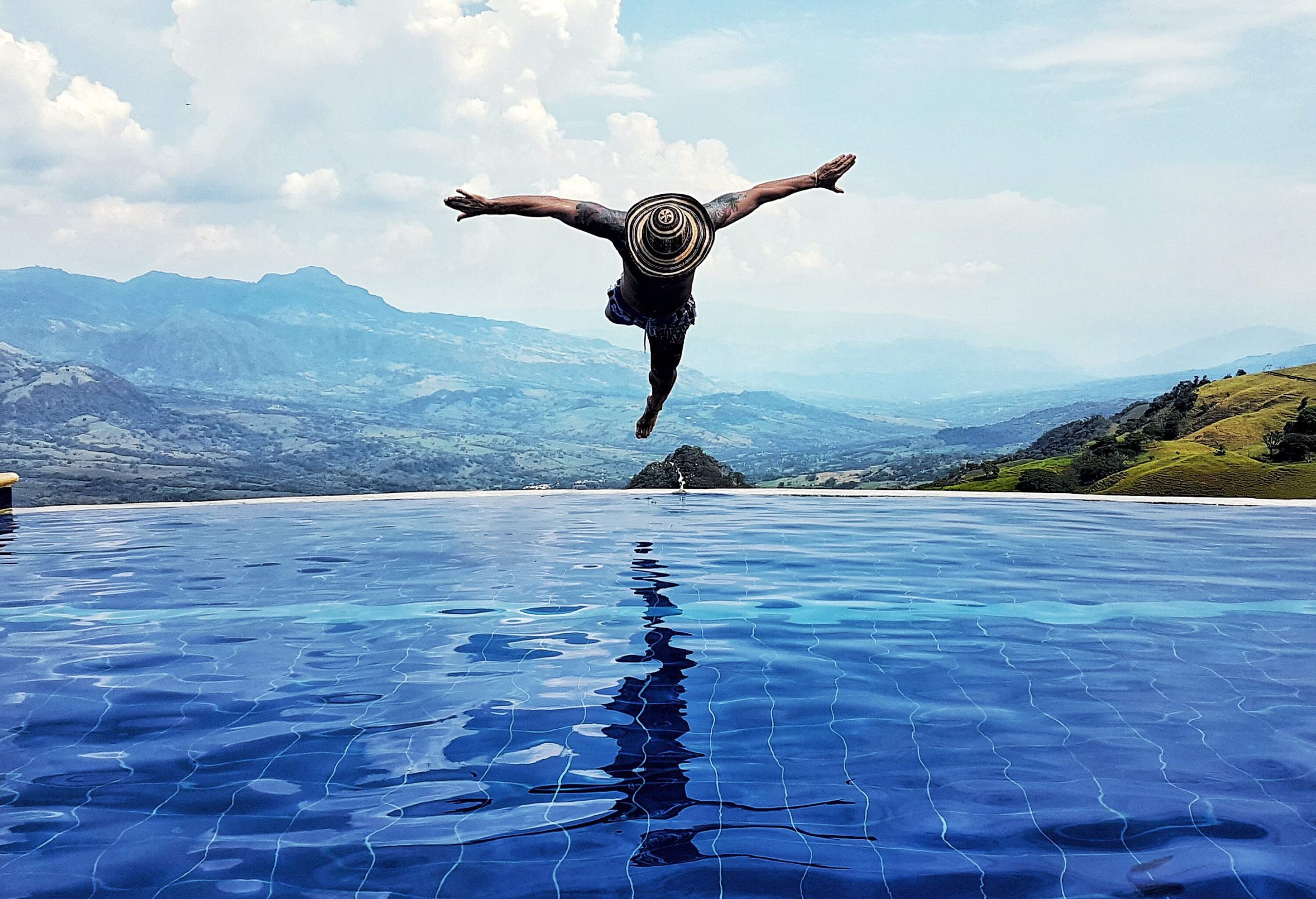 A person in a sun hat dives into an inviting infinity pool with a picturesque mountainous landscape in the background.