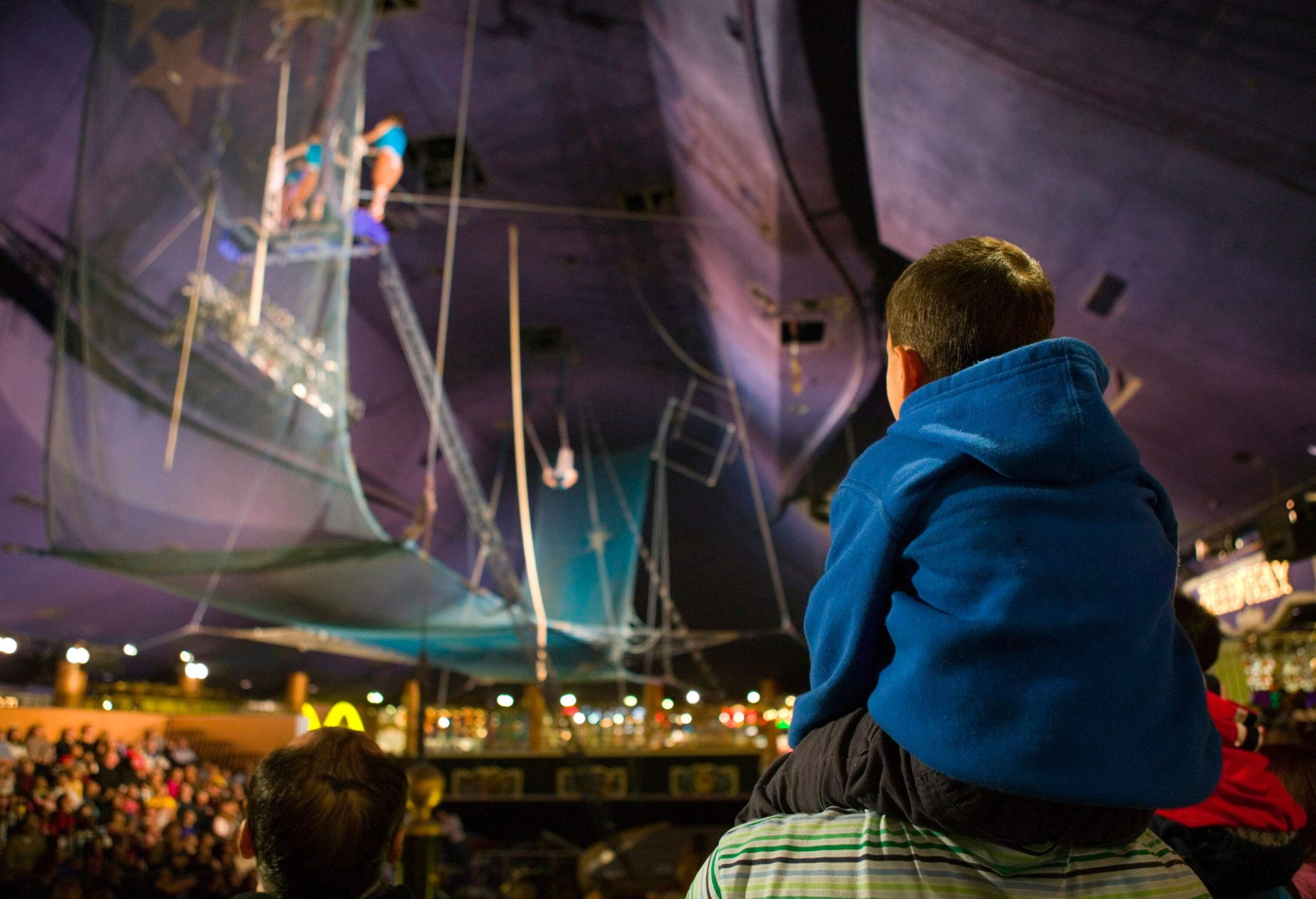 A child perches upon someone's shoulder, their eyes wide with wonder, amid the captivating spectacle of a vibrant circus show.