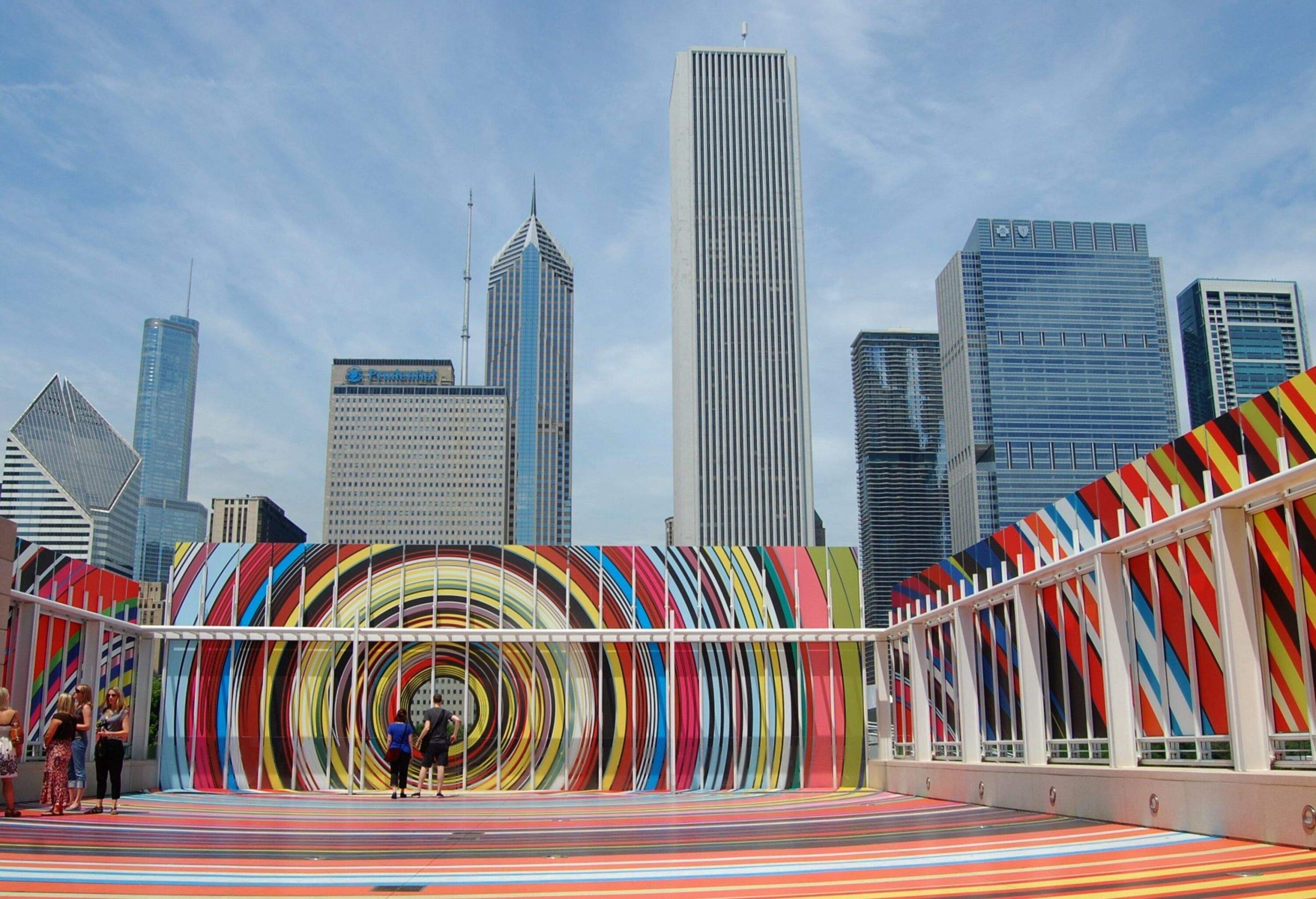 People in an open area with colourful spiral art against the modern towering skyscrapers in the background.