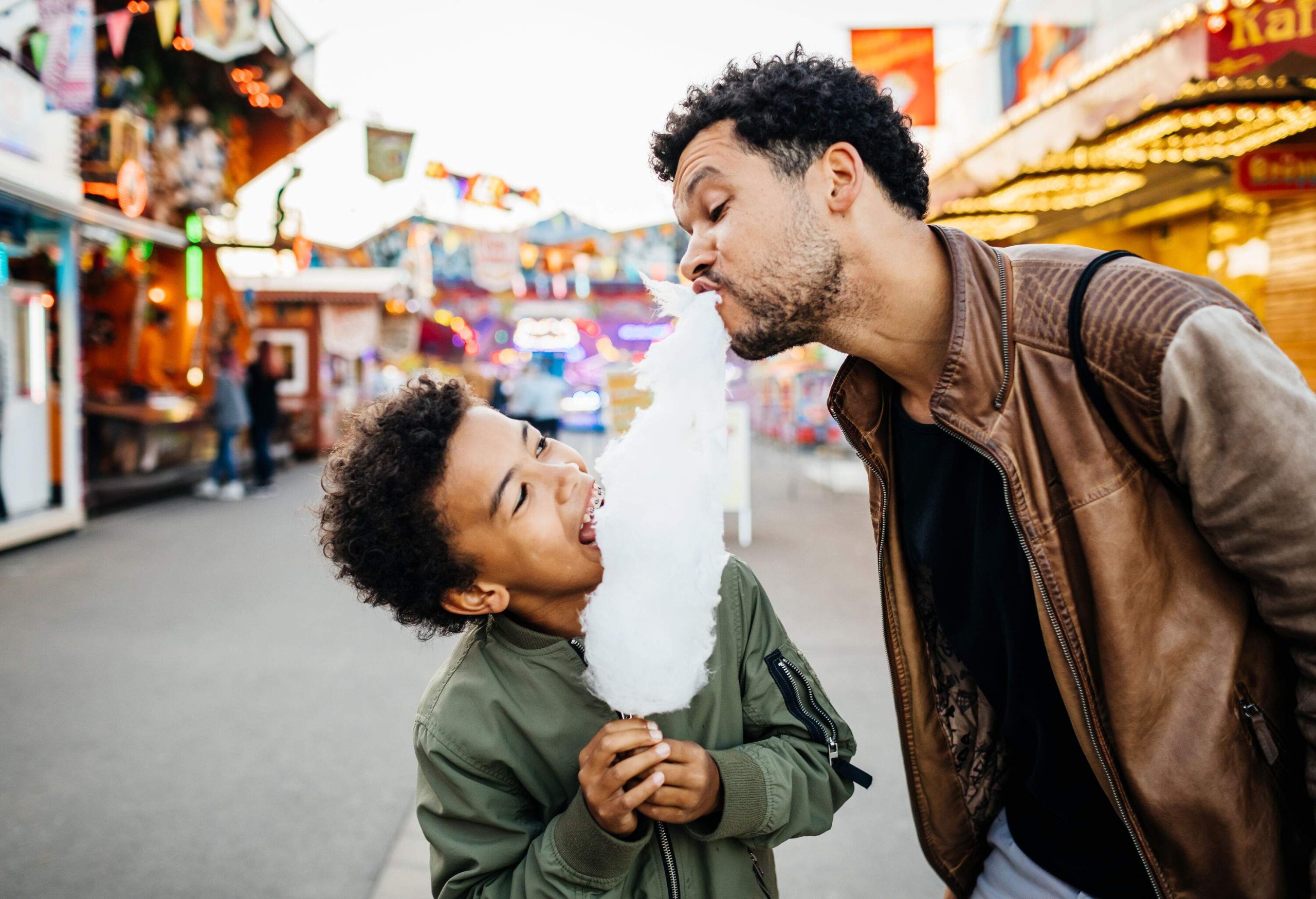 A happy father and son sharing a cotton candy on a stick against the brightly lit amusement park.
