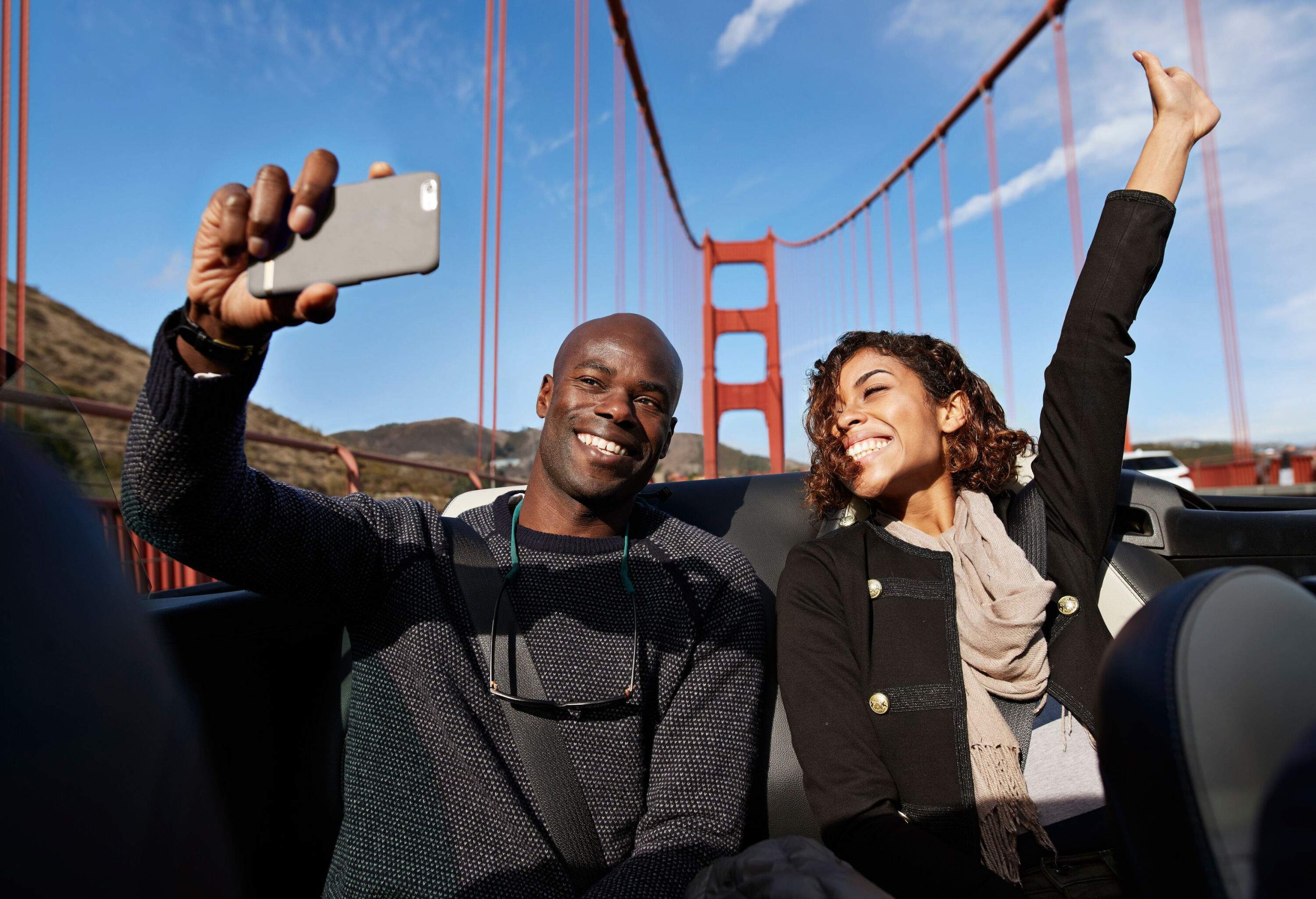 A man and a woman taking a selfie as they ride in the backseat of a car with the top down on a bridge.