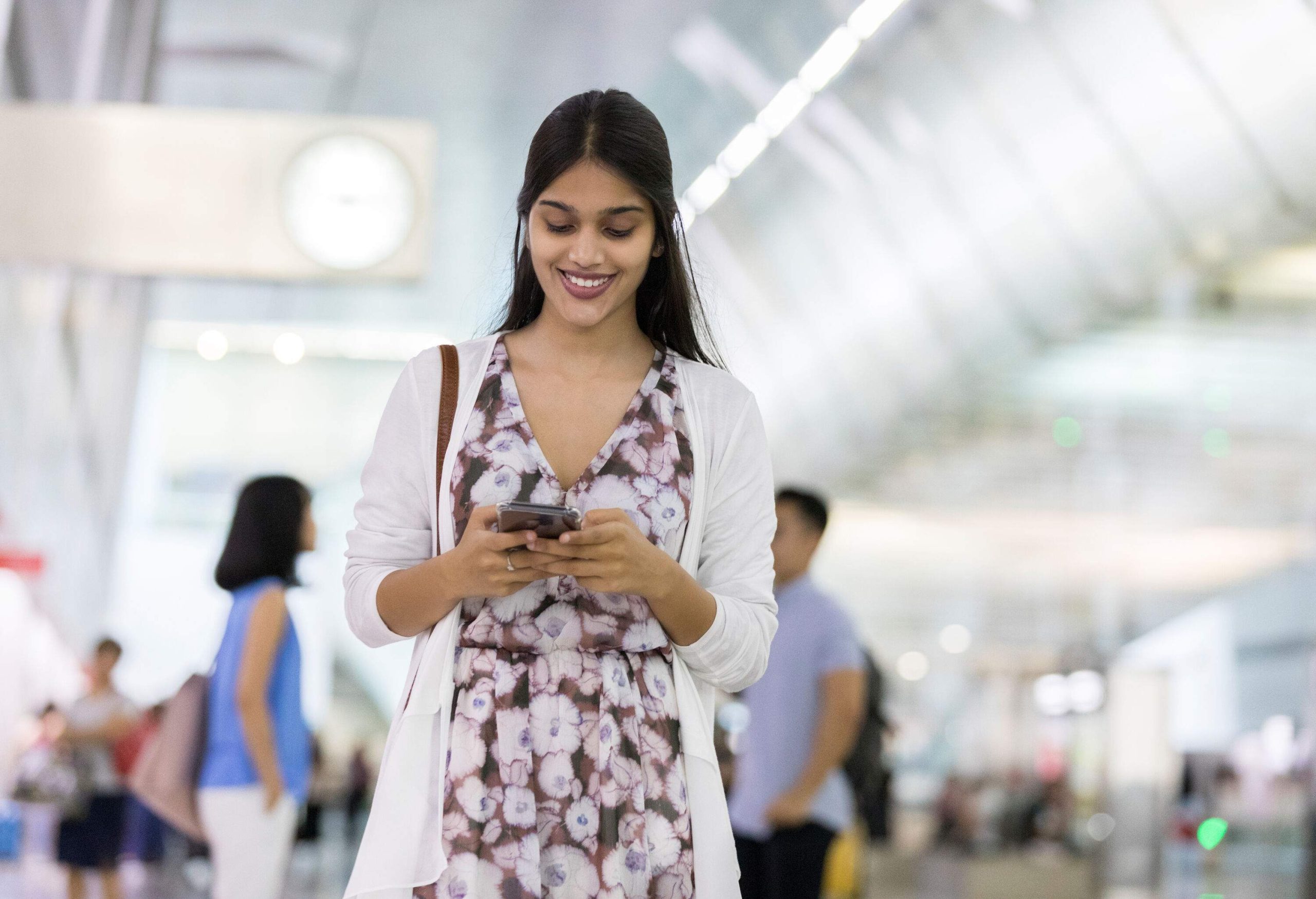 A raven-haired woman wearing a white cardigan over a nice dress checking on her phone while standing at an airport.