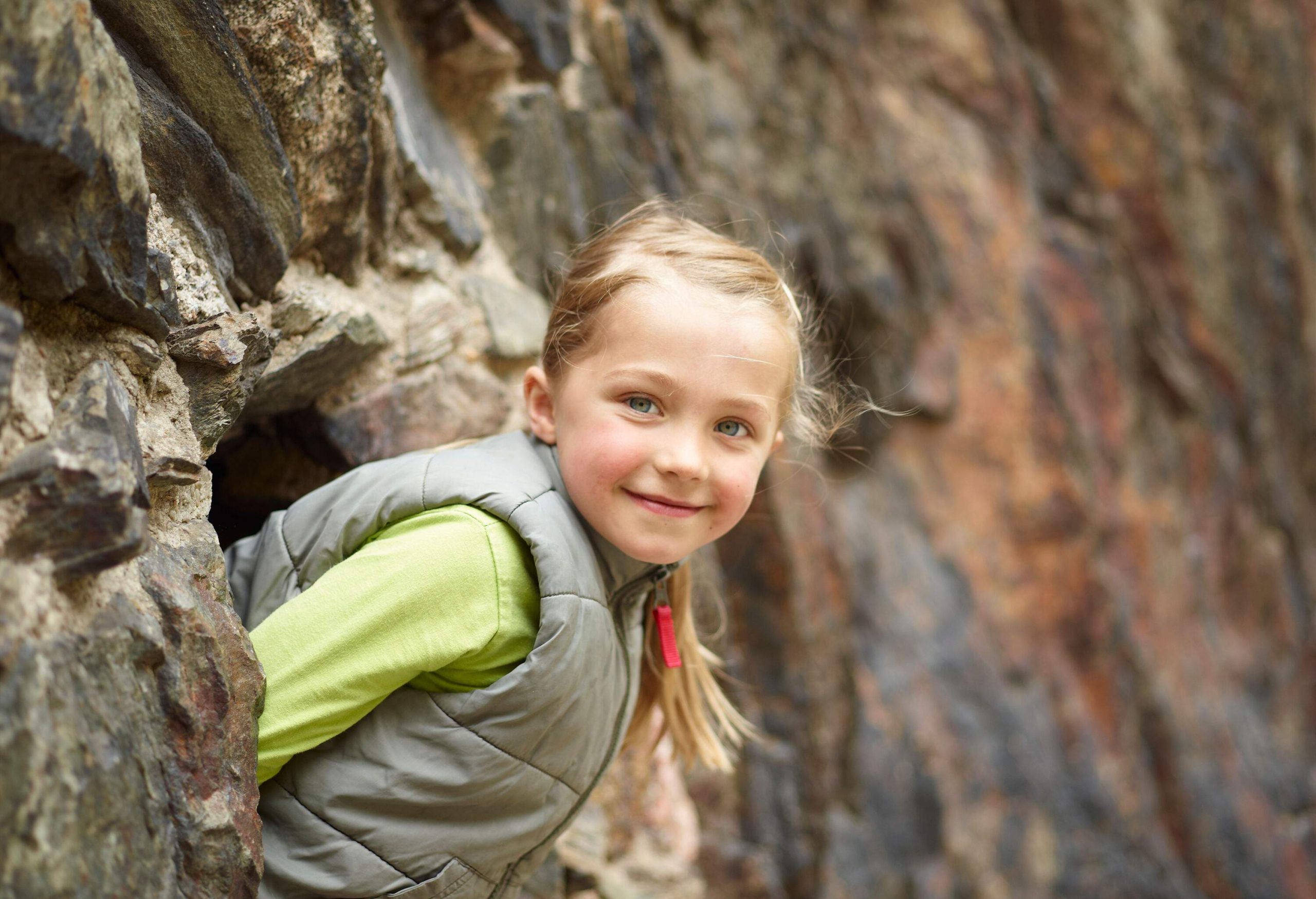A happy little girl peeking out from a cave in a mountain and looking outside.