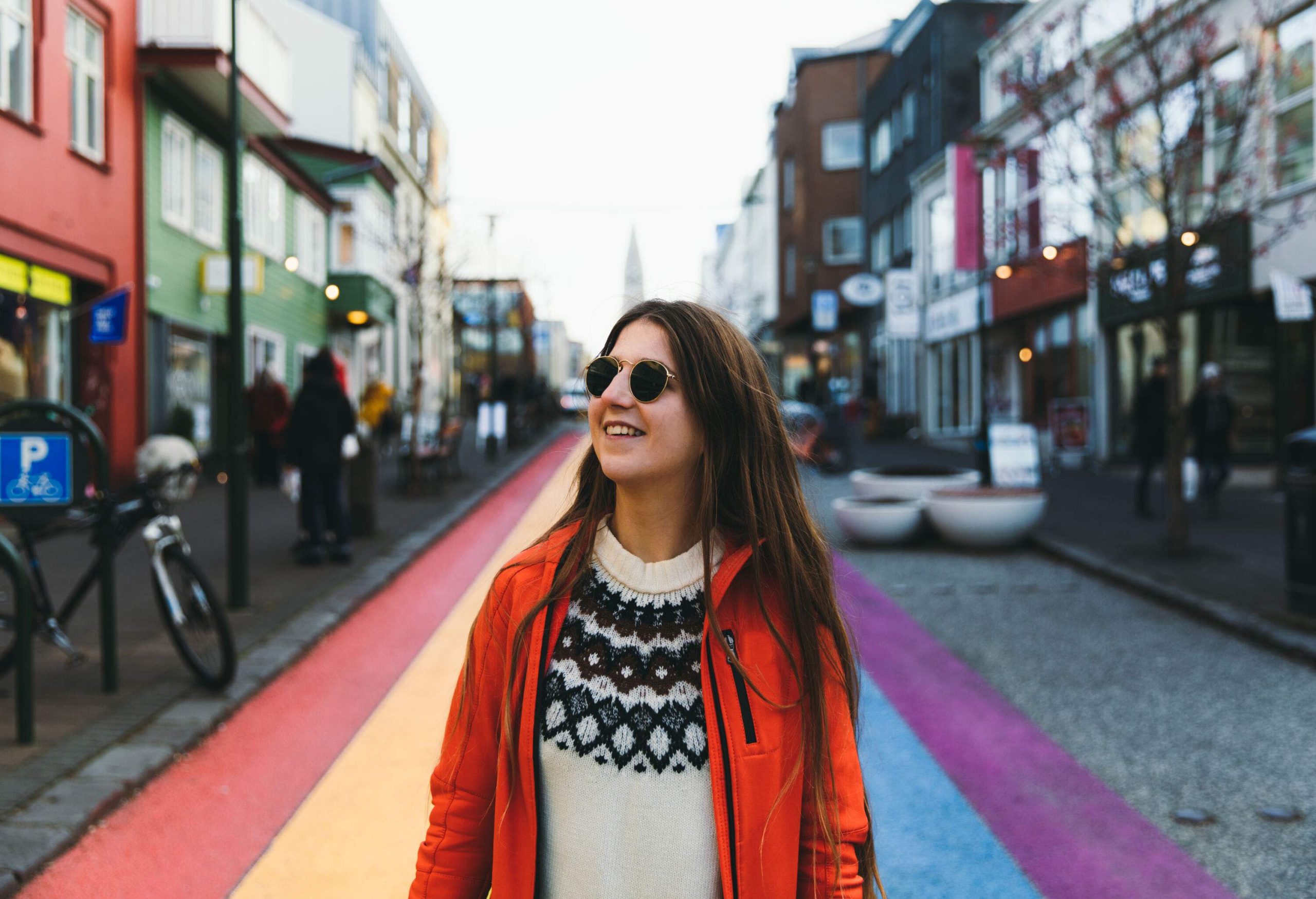 A long-haired woman walking through the colourful streets, wearing a red jacket, knitted sweater, and sunglasses.
