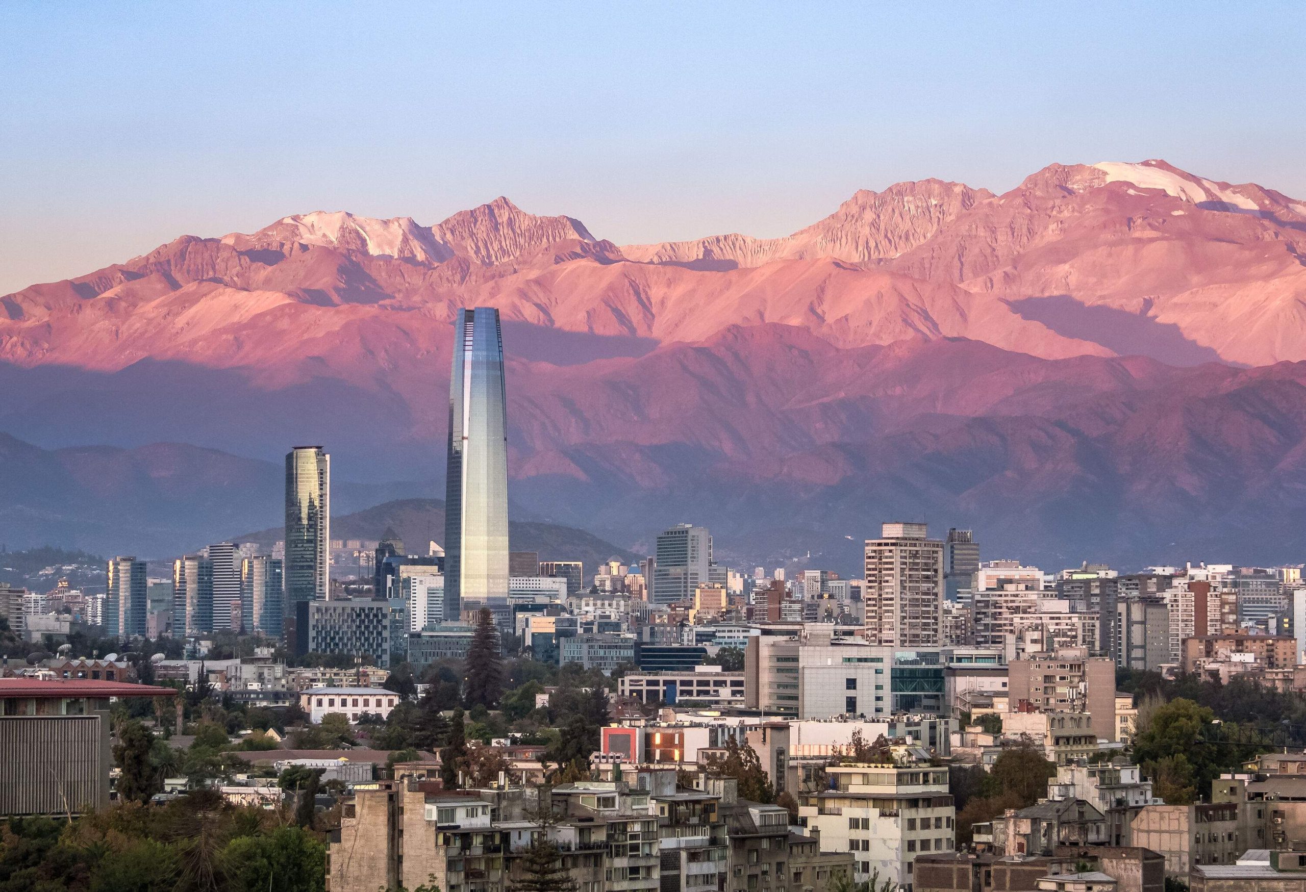 Gran Torre Santiago, a 62-storey skyscraper, dominates the cityscape at the base of tall bald sunlit mountains.