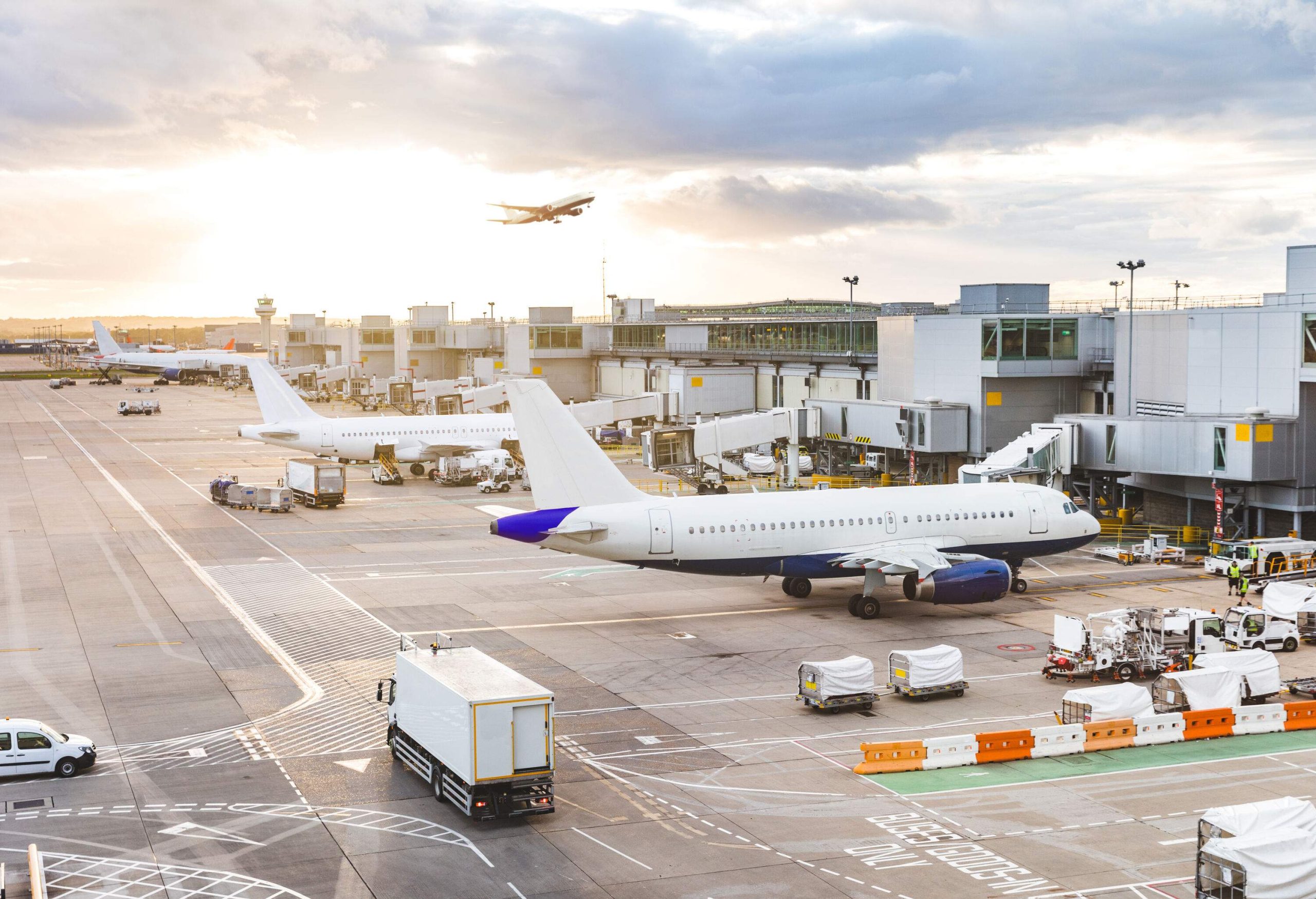 A busy airport with parked aeroplanes and service vehicles in the flight line against the sunset sky.